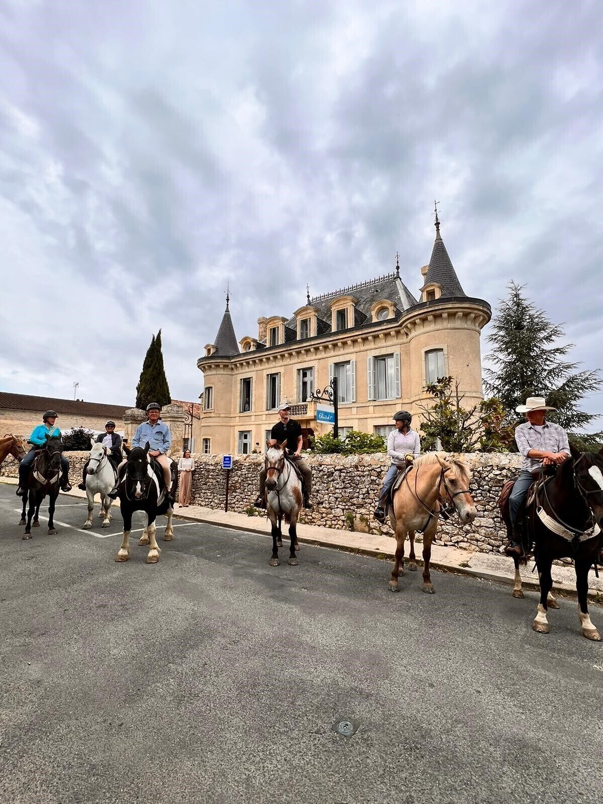 A group of people riding horses in front of a large, historic building with multiple towers and a stone wall.