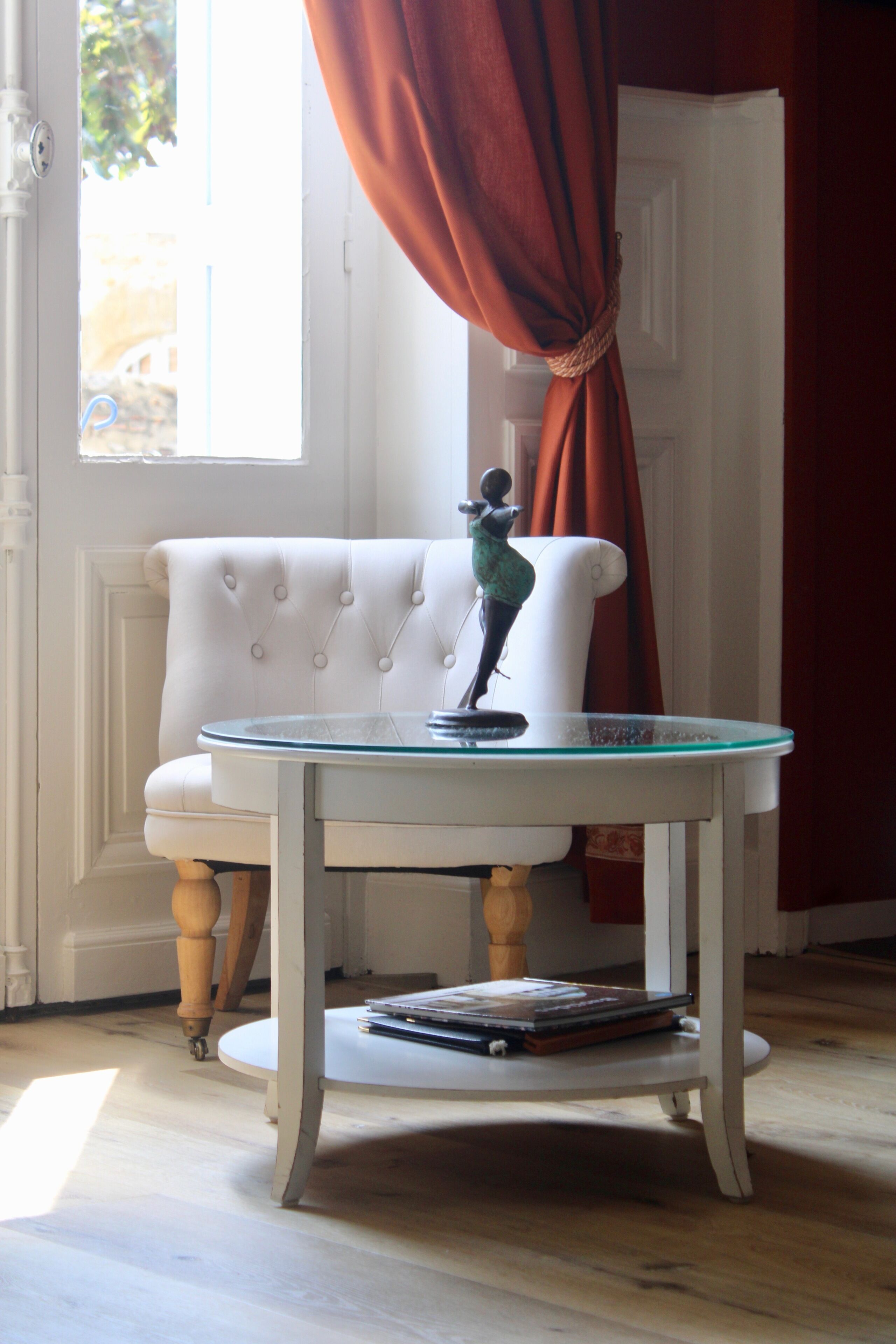 A white upholstered chair with a glass table, a small statue, and a window with curtains.