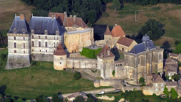 Vue aérienne d’un ensemble de bâtiments historiques formant un château, comprenant plusieurs constructions, dont un grand bâtiment central surmonté d’une tour, le tout entouré de bâtiments plus petits et de murs fortifiés.