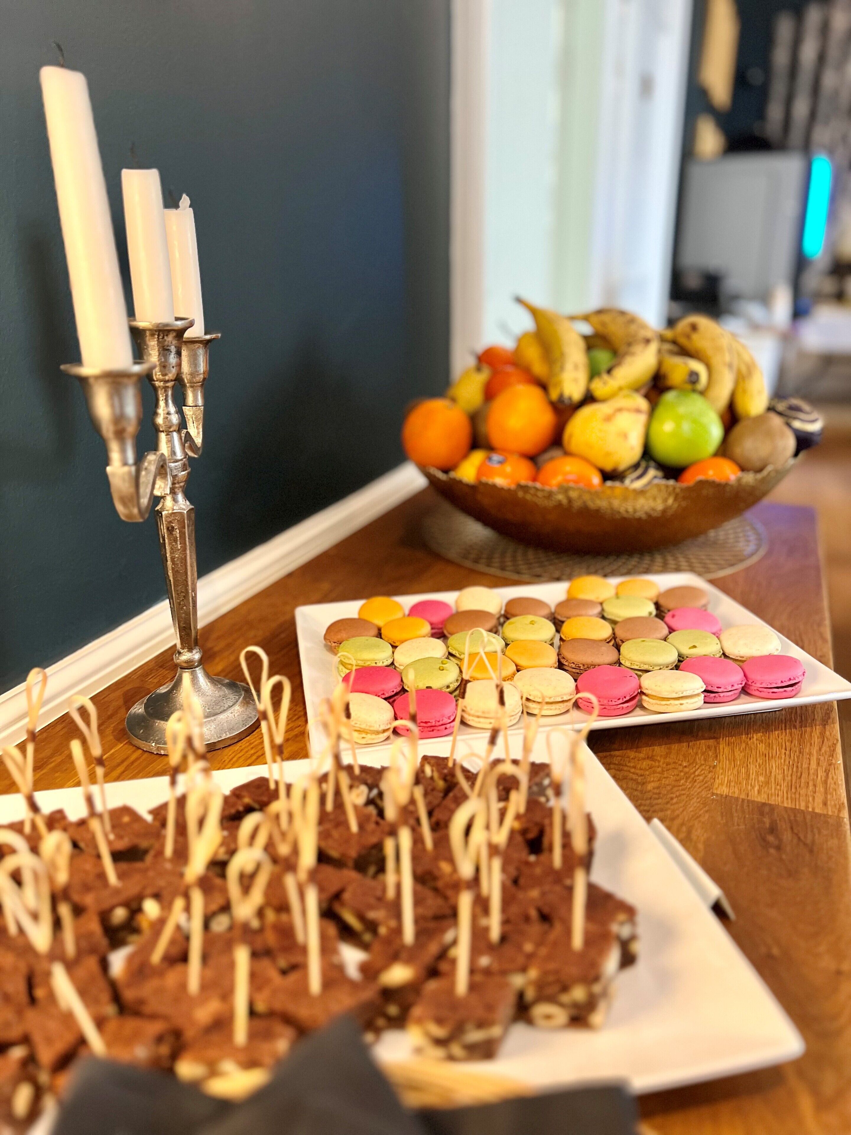Una mesa con varios postres, incluyendo macarons y brownies, y un frutero al fondo.