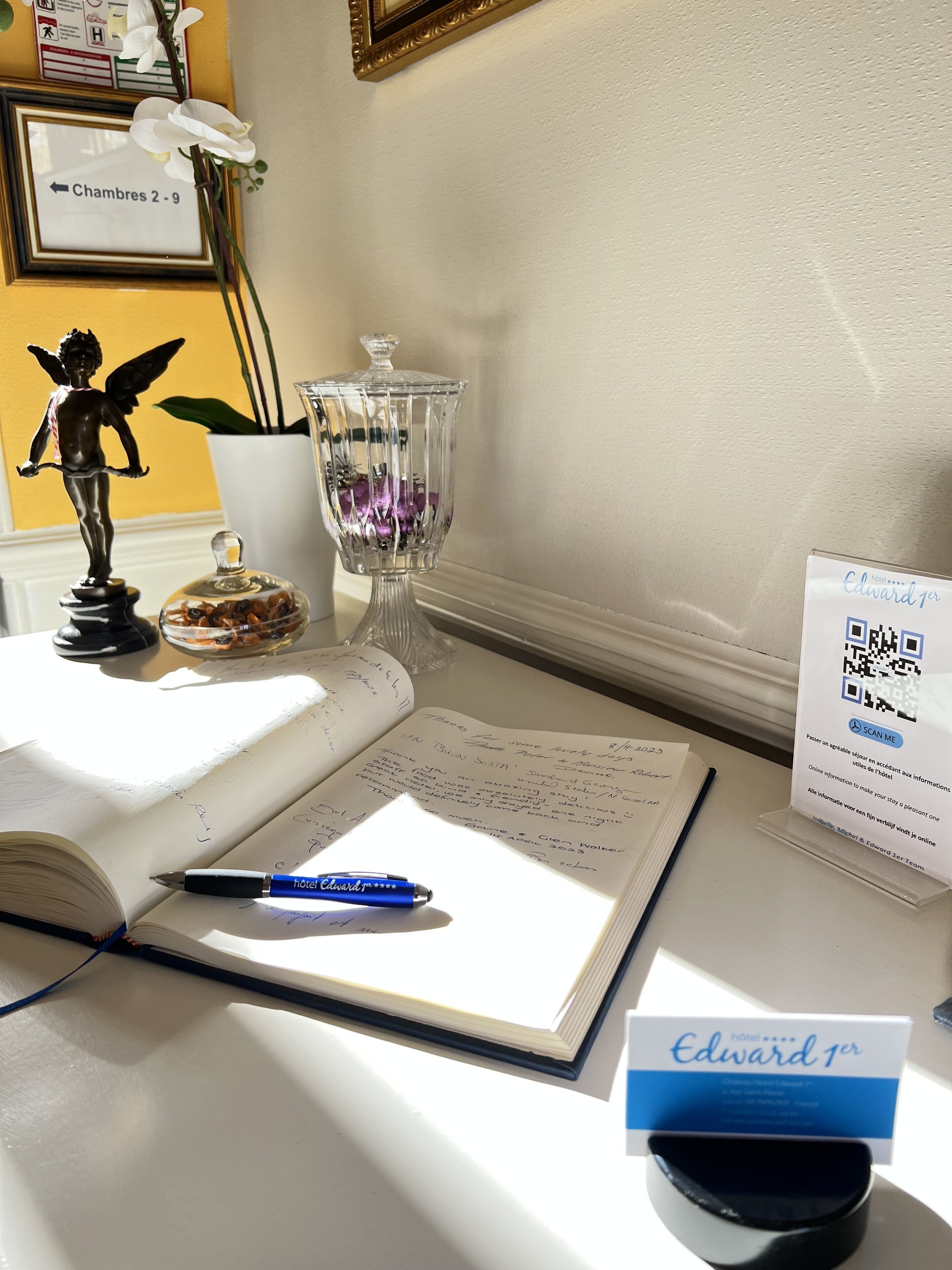 A hotel room with a bed, a bedside table with a book, a pen, a decorative angel figurine, a glass container with flowers, and a framed sign indicating room numbers.