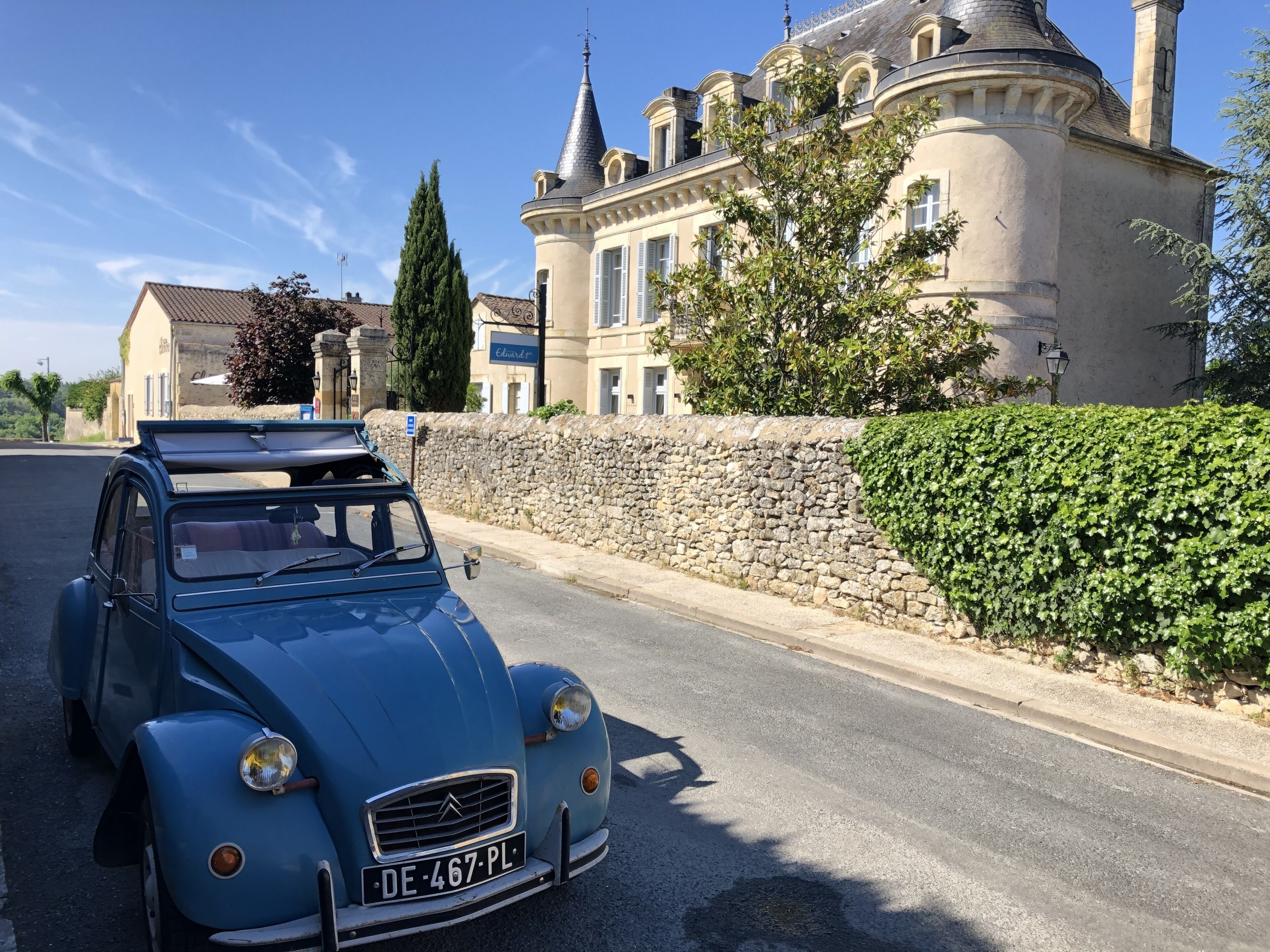 A vintage blue car parked on a street with a stone wall and a large building with a turret in the background.