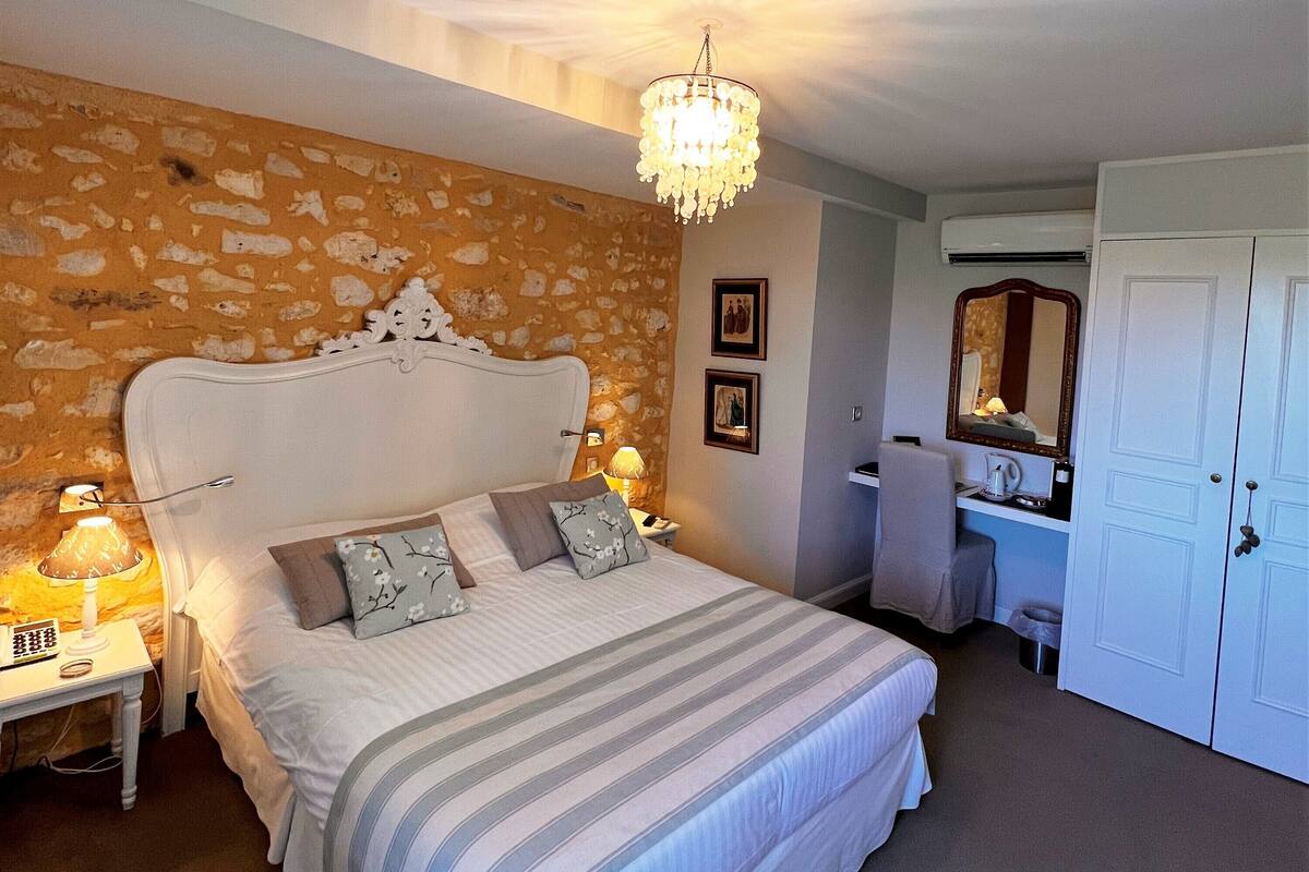 A bedroom with a stone wall, a large bed with a chandelier, a desk with a lamp, and a wardrobe.