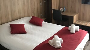 Ein Hotelzimmer mit einem großen Bett, zwei roten Kissen, einem hölzernen Kopfteil, einem Schreibtisch mit Stuhl, einem an der Wand montierten Fernseher und einer kleinen Ablage mit Toilettenartikeln.