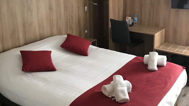 A hotel room with a large bed, two red pillows, a wooden headboard, a desk with a chair, a TV mounted on the wall, and a small shelf with toiletries. - Kyriad - Narbonne Sud (Narbonne)