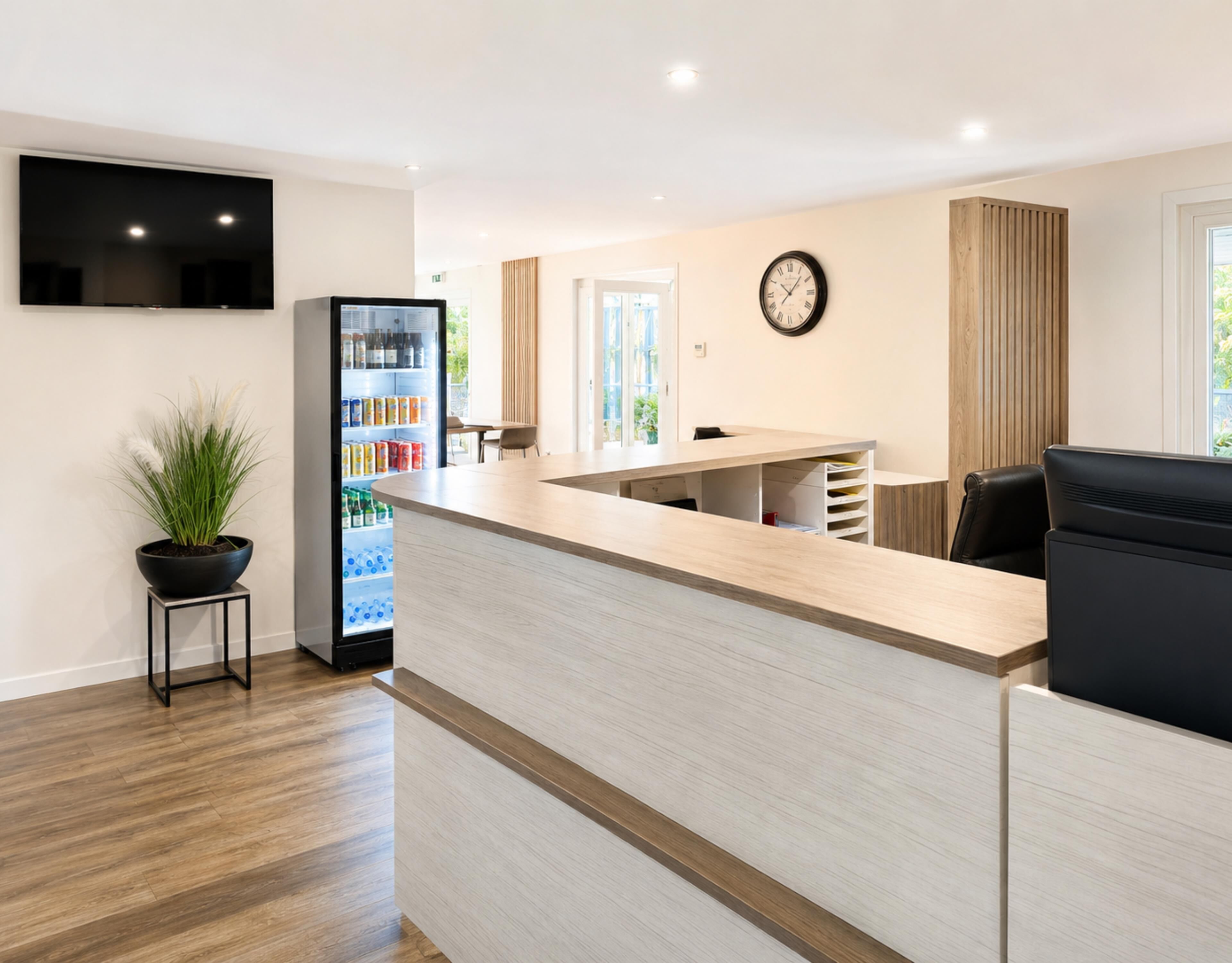 Un espace d’accueil comprenant un comptoir, un téléviseur fixé au mur, une horloge et un réfrigérateur rempli de boissons.