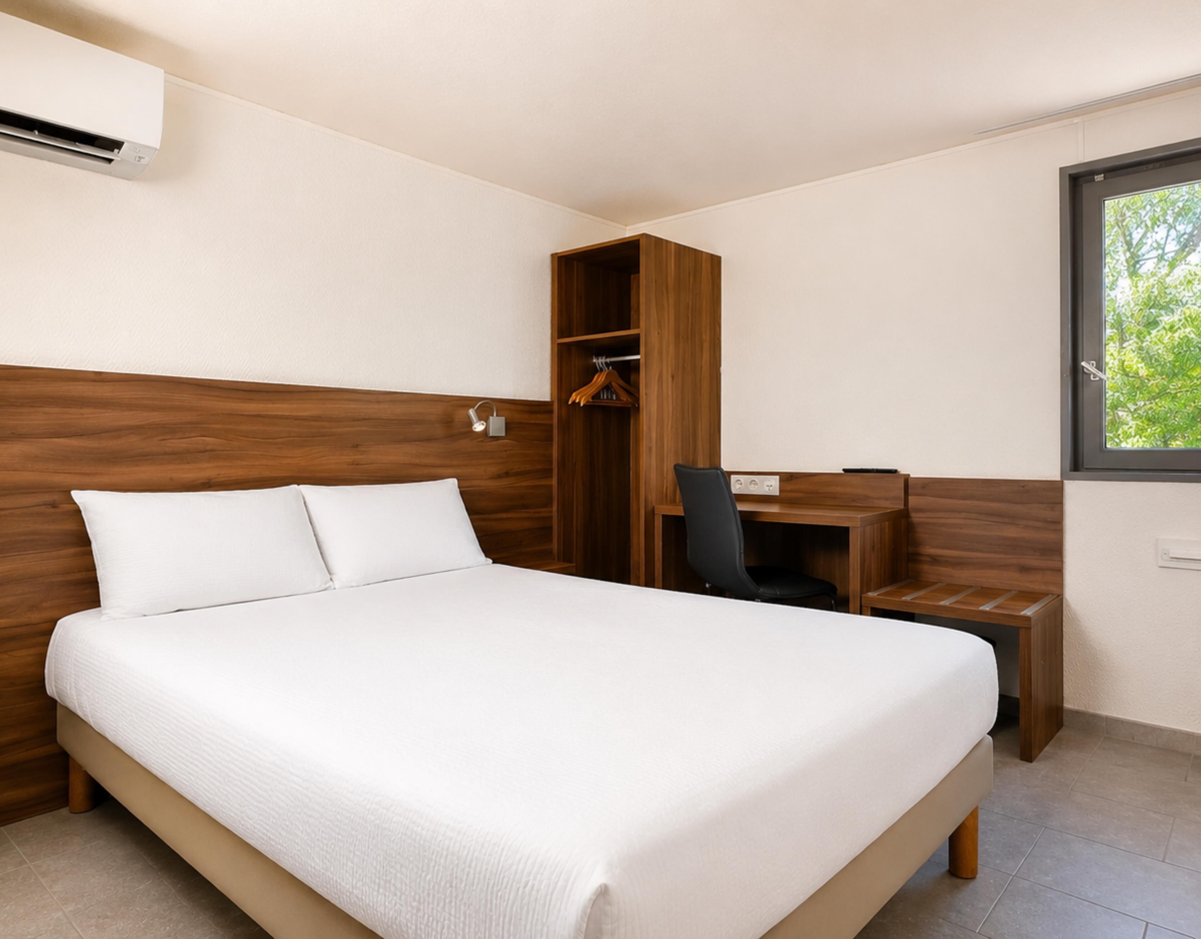 Ein Hotelzimmer mit einem großen Bett, einem Holzschrank, einem Schreibtisch mit Stuhl und einem Fenster mit Blick auf Bäume.