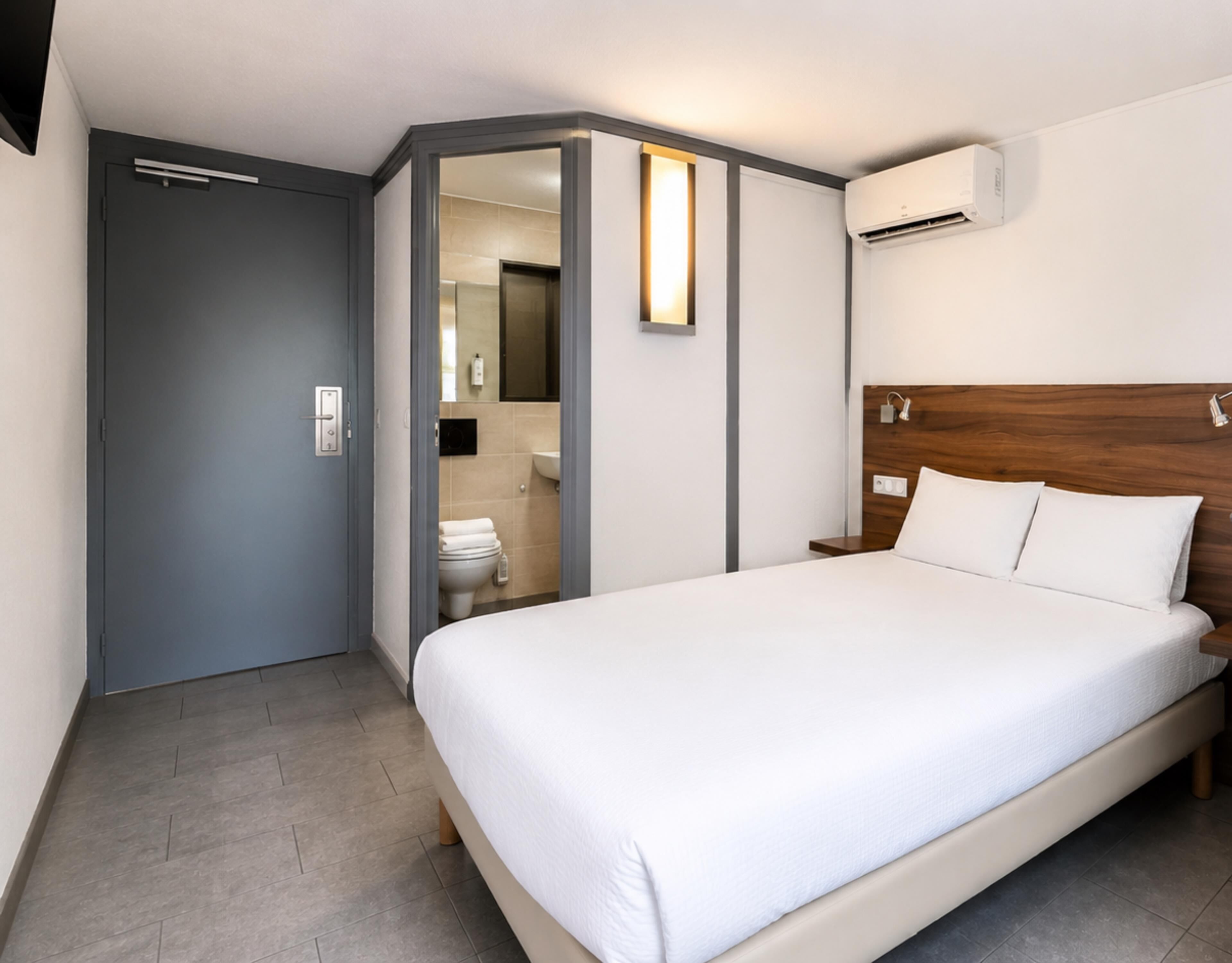 A hotel room with a bed, a door leading to a bathroom, and an air conditioning unit on the wall.