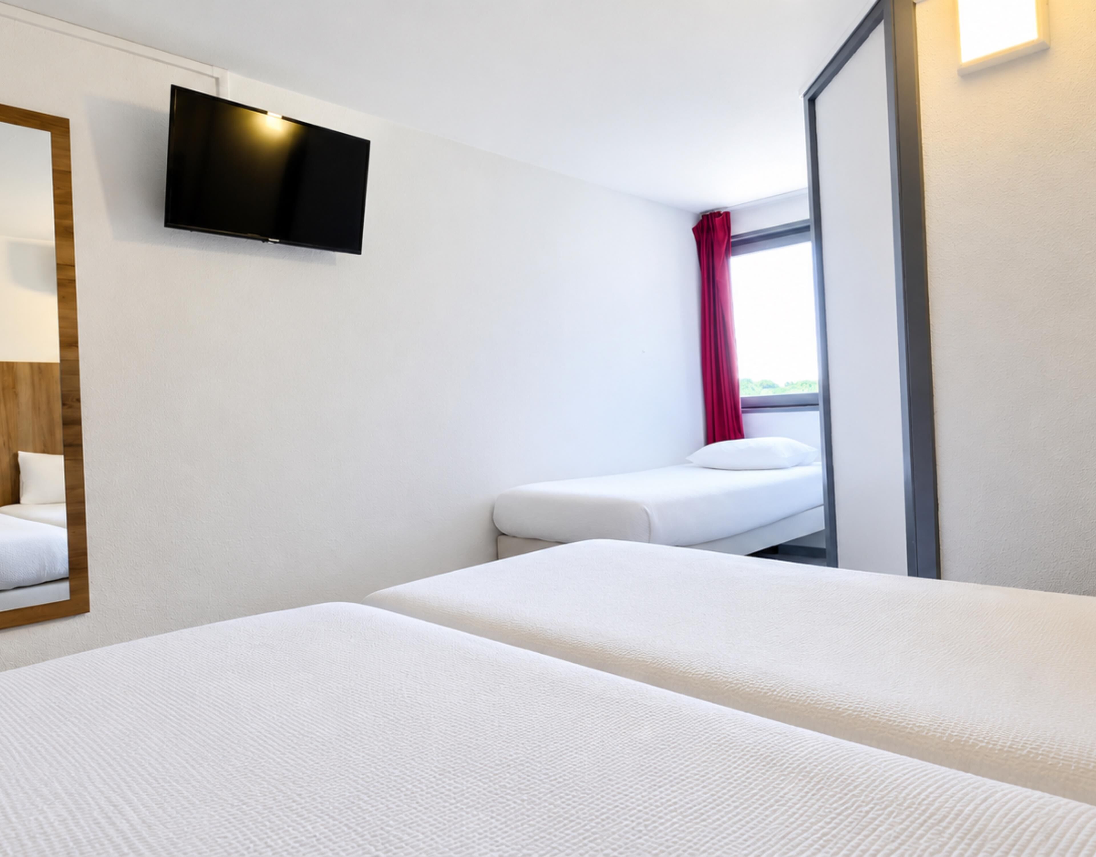 A hotel room with two beds, a TV mounted on the wall, and a window with a red curtain.