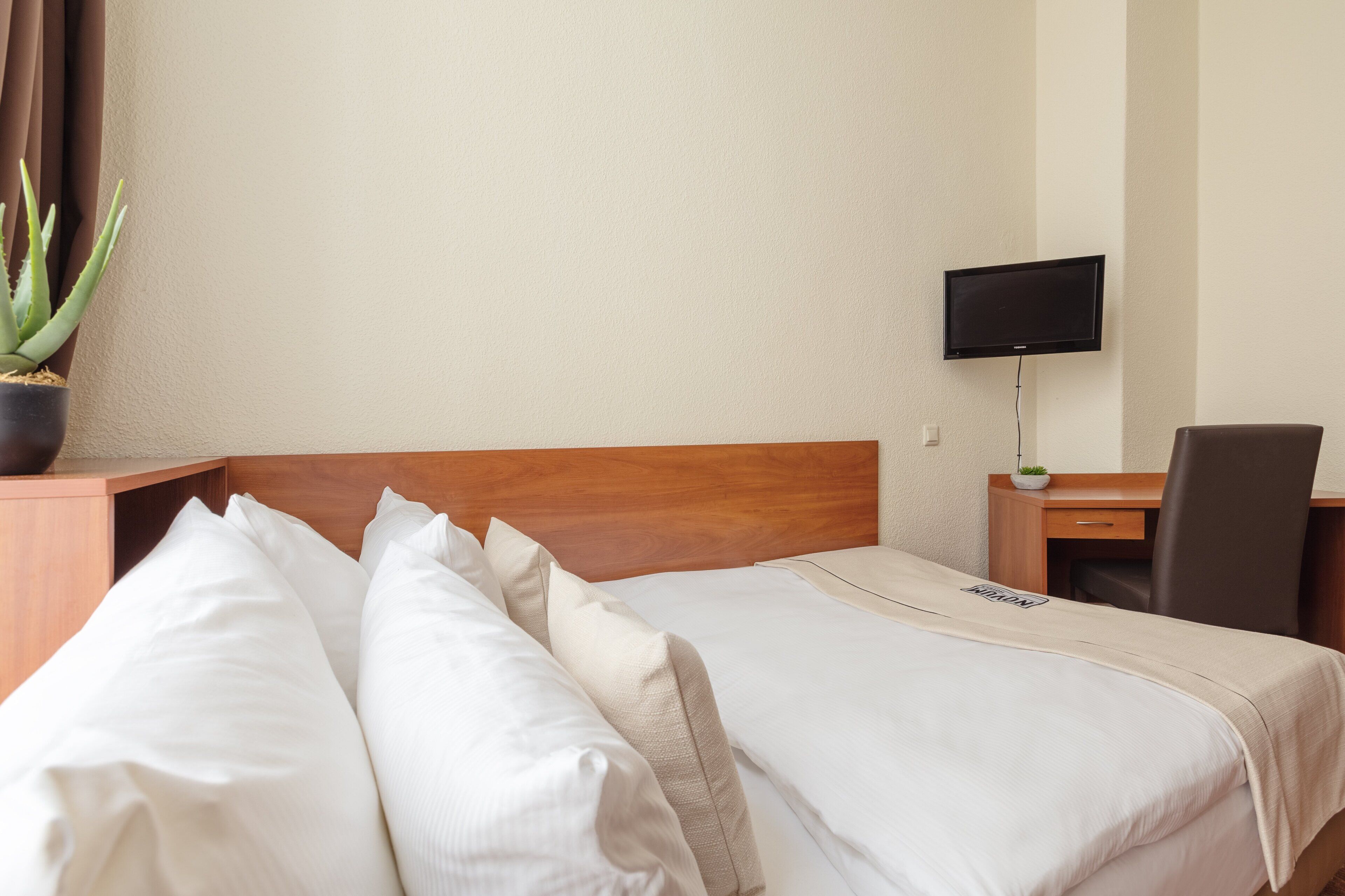 A hotel room with a wooden headboard, a flat-screen TV mounted on the wall, a desk with a chair, and a potted plant.