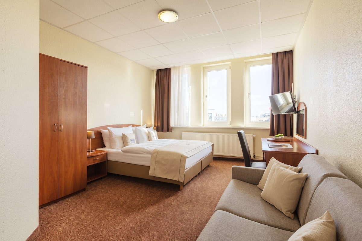 Une chambre d’hôtel comprenant un lit, un canapé, un bureau avec une lampe, une armoire et une fenêtre avec des rideaux.