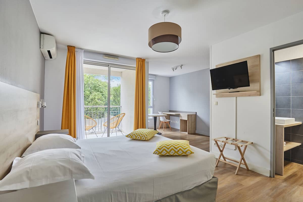 Una habitación de hotel con cama, escritorio, televisor y balcón con cortinas amarillas.