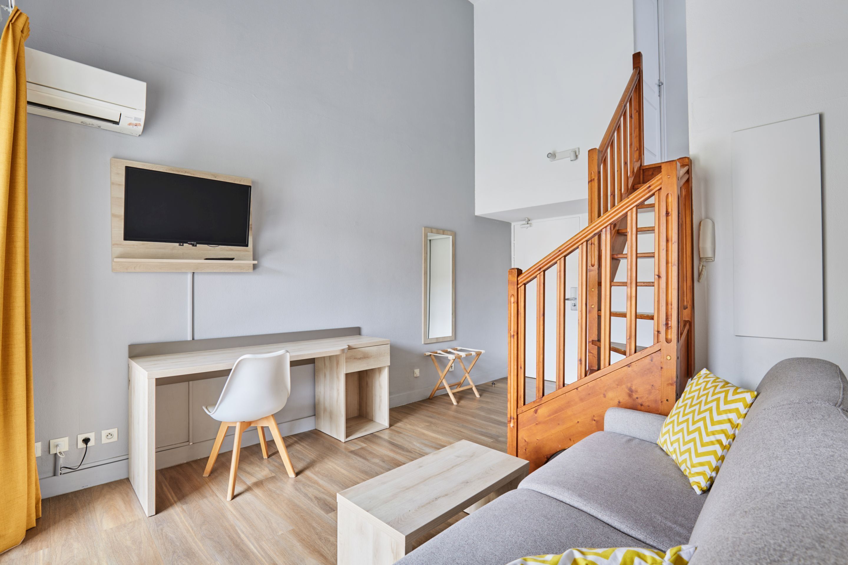 Een moderne woonkamer met een houten bureau, een grijze bank, een houten trap en een wandtelevisie.