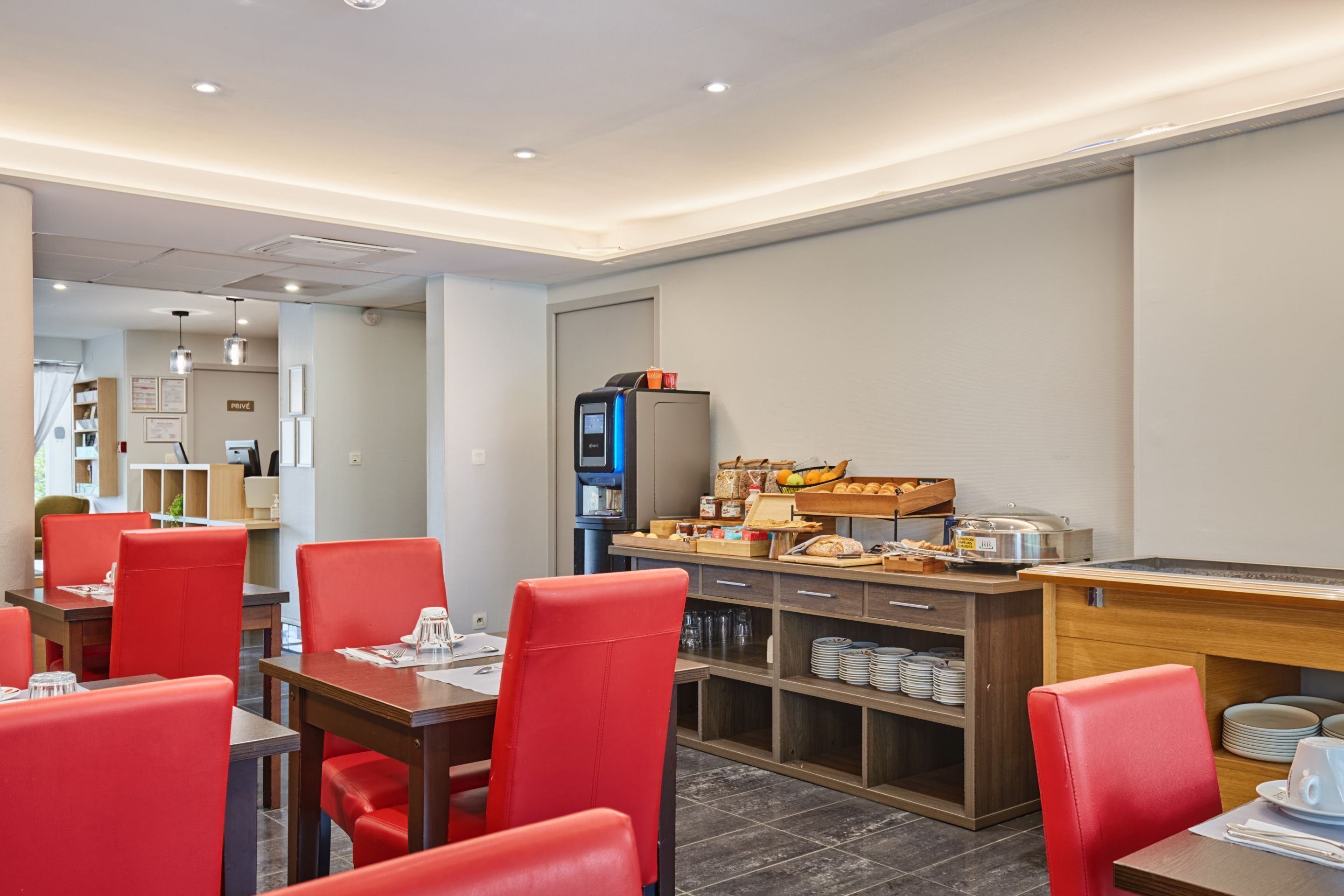 Un espace repas moderne avec des chaises et des tables rouges, un buffet proposant des plats et des boissons, et un téléviseur.