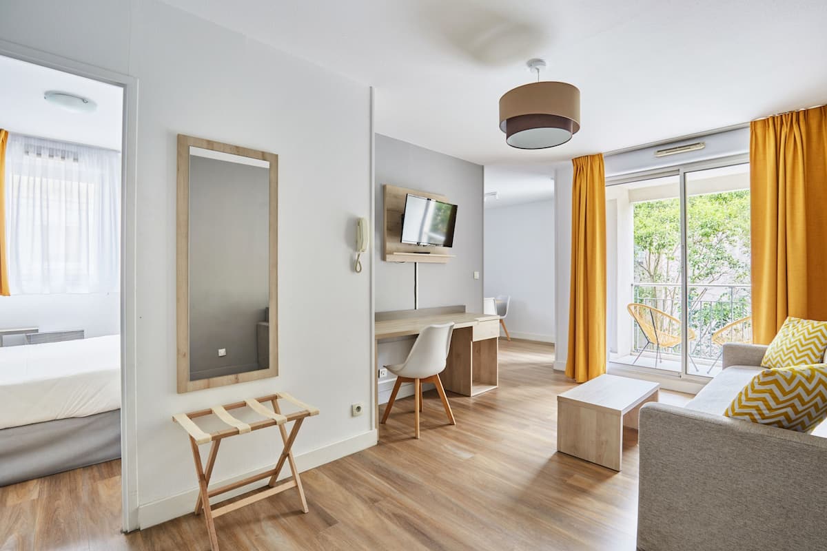 Habitación de hotel moderna con suelo de madera, una cama, un escritorio con una silla, un sofá, un televisor y un balcón con cortinas amarillas.
