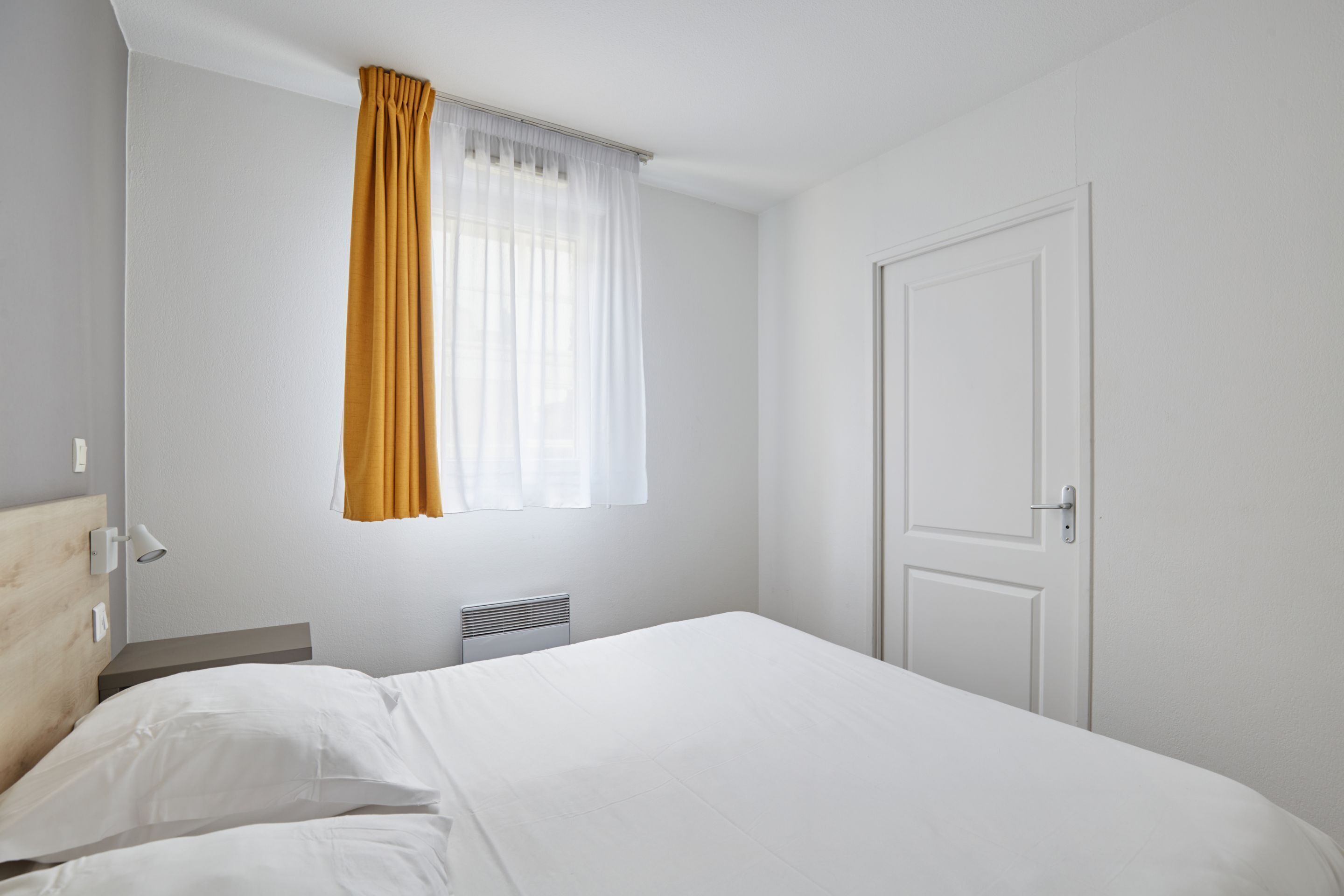 A hotel room with a bed, a window with curtains, a door, and a wall-mounted light.