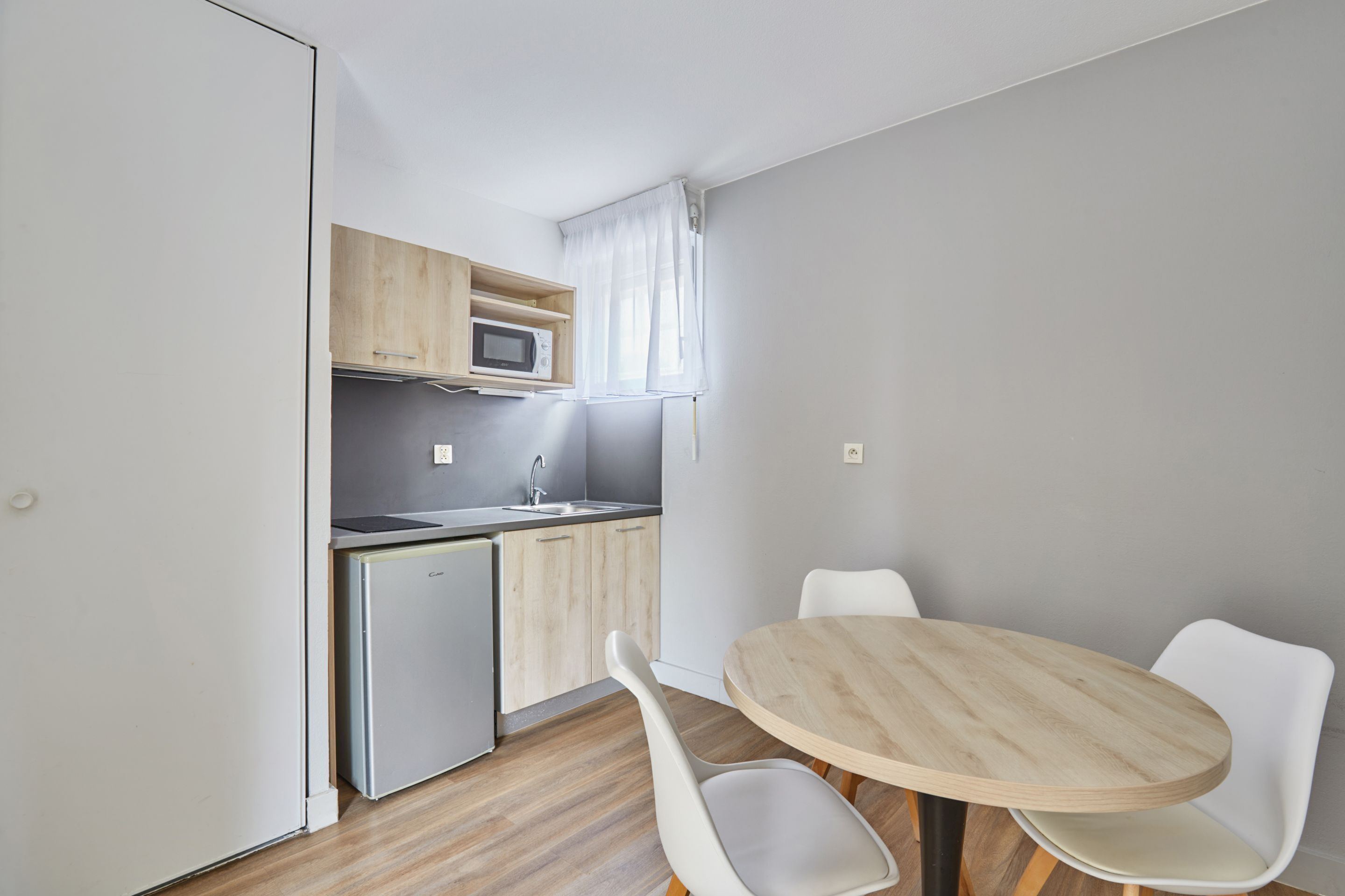 Cocina compacta con microondas, lavamanos y armarios de madera. Hay una mesa redonda de madera con dos sillas blancas.