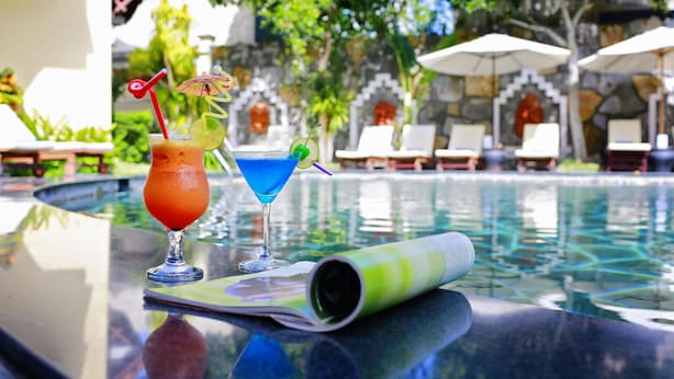Une scène au bord de la piscine, avec deux boissons sur une table, un menu enroulé et des chaises longues en arrière-plan.