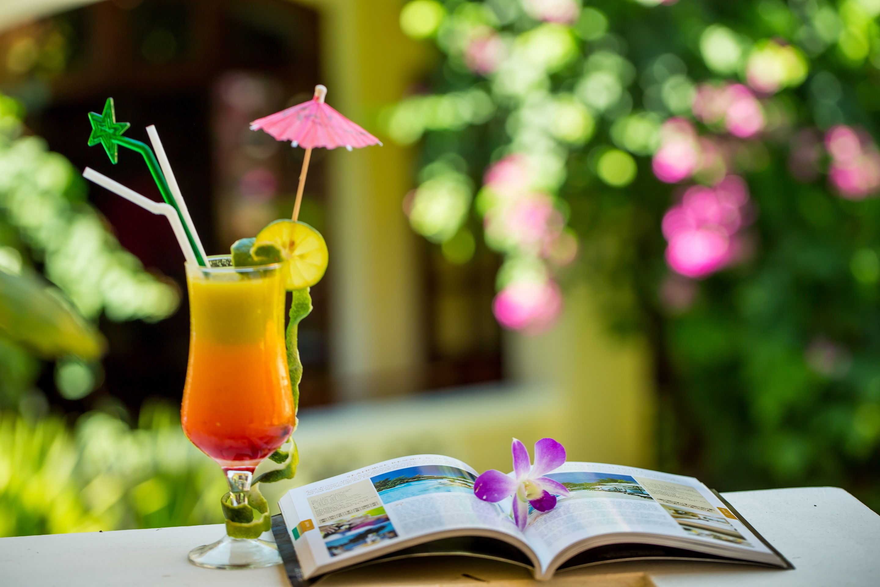 A glass of orange juice with a star-shaped straw and a slice of lime, an open magazine, and a pink umbrella on a table.