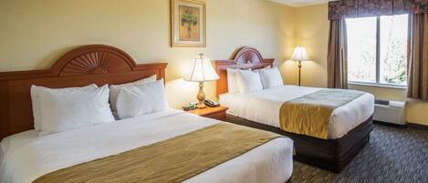 Habitación de hotel con dos camas, cada una con cabecero de madera, ropa de cama blanca y una manta marrón. Hay un cuadro en la pared, una ventana con cortinas y una lámpara en cada mesita de noche.