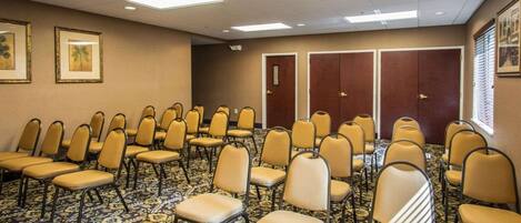Un salón de conferencias con filas de sillas, una alfombra estampada, luces de techo y puertas.