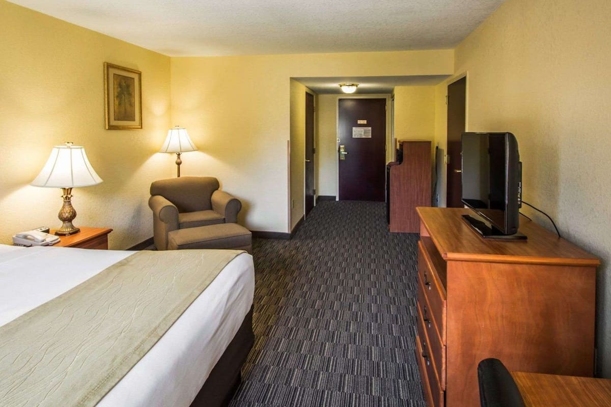A hotel room with a bed, two lamps, a chair, a desk with a television, and a door leading to another room.