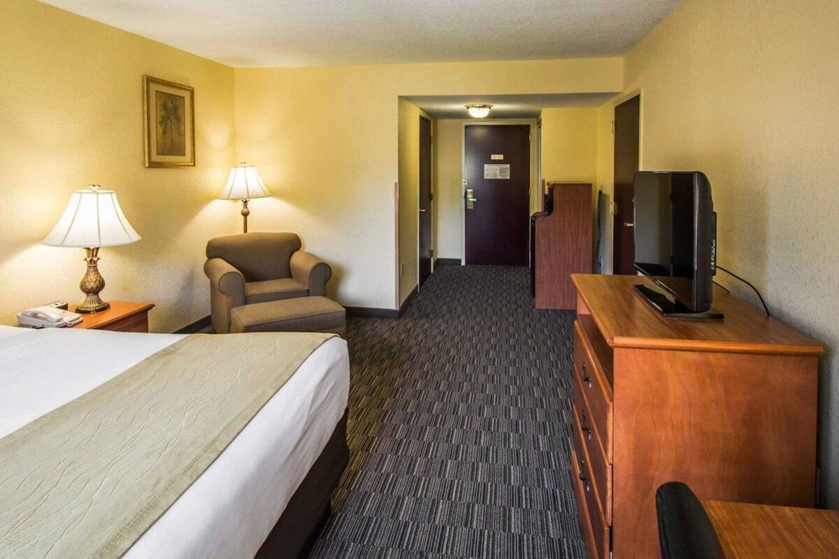 A hotel room with a bed, two lamps, a chair, a desk with a television, and a door leading to another room.