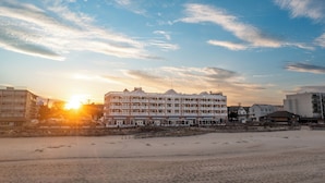 Un hotel frente a la playa con varios balcones, una playa arenosa en primer plano y un cielo despejado con nubes.