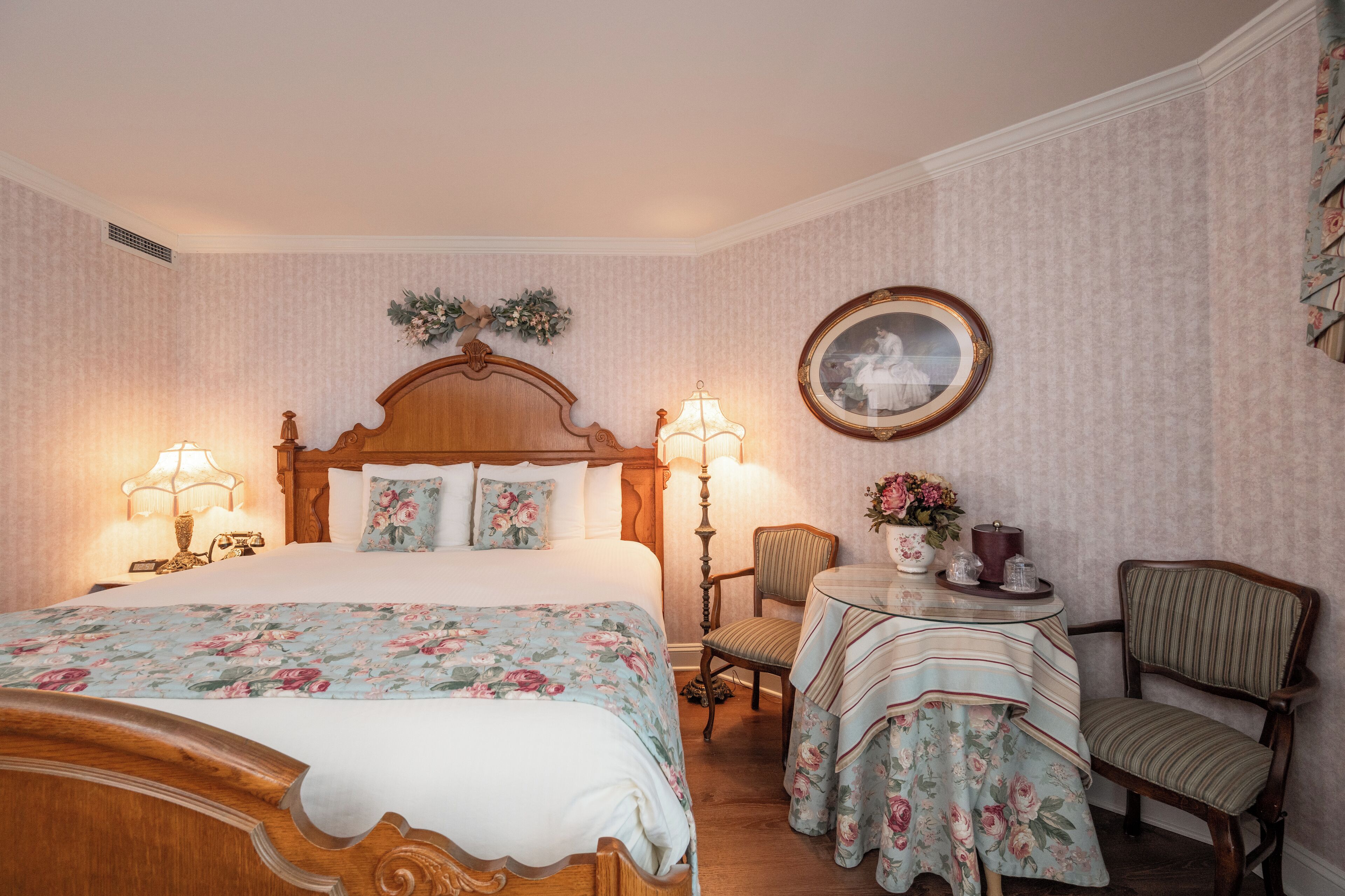 Un dormitorio ordenado con una cama grande, dos sillas, una mesita redonda y un cuadro en la pared.