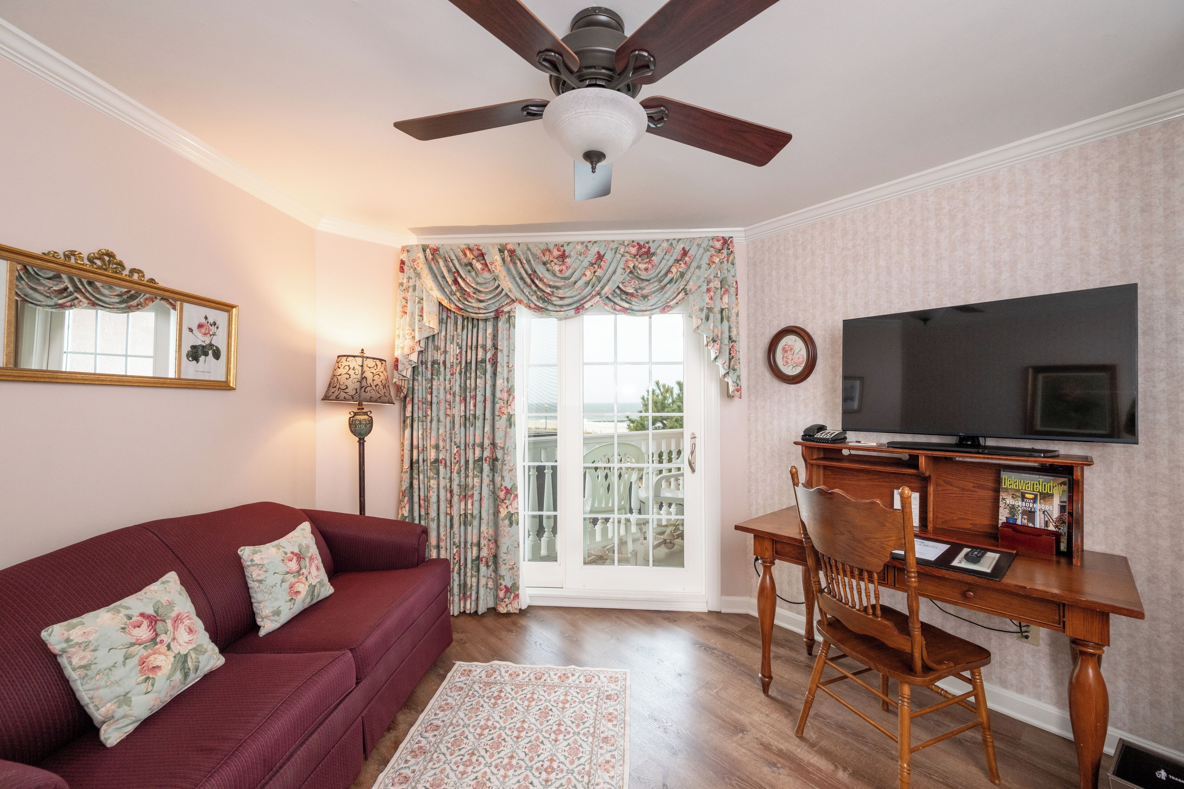 A living room with a red sofa, floral curtains, a ceiling fan, a television, and a wooden desk.