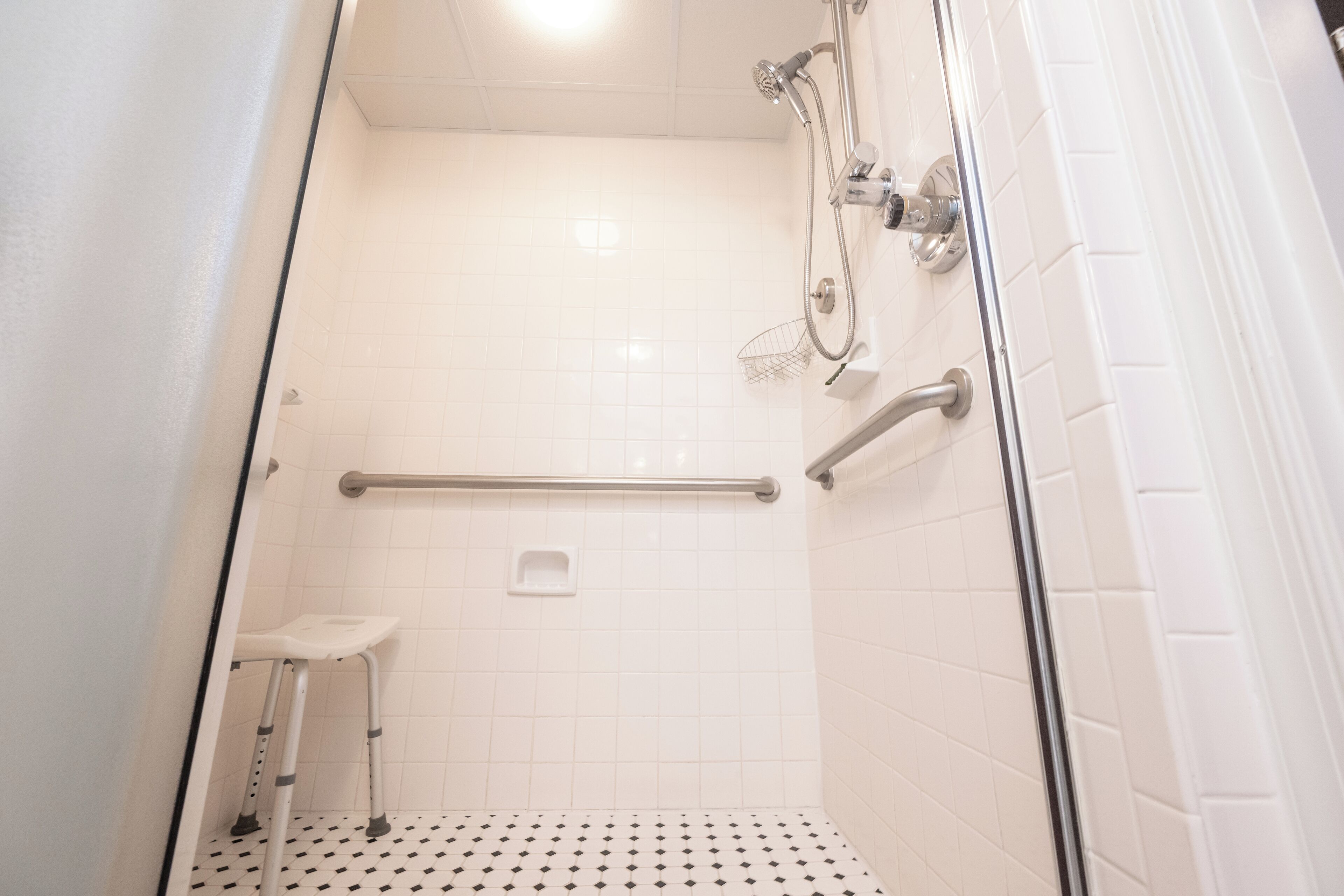 A small, enclosed shower area with white tiled walls and floor, a handheld showerhead, a handheld showerhead, a grab bar, and a small white stool.