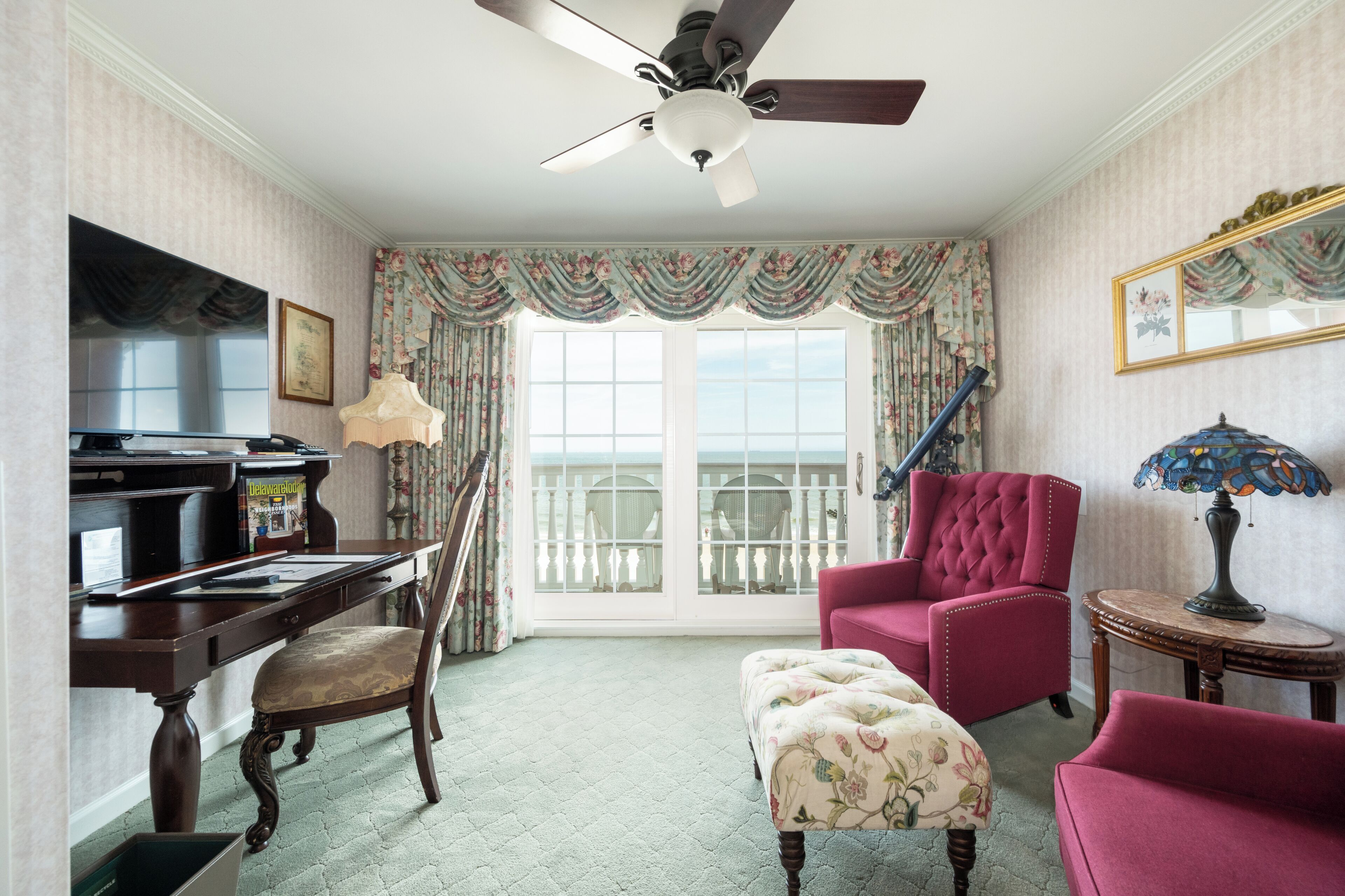 A room with a wooden desk, a red armchair, a matching ottoman, a ceiling fan, and a large window with curtains.