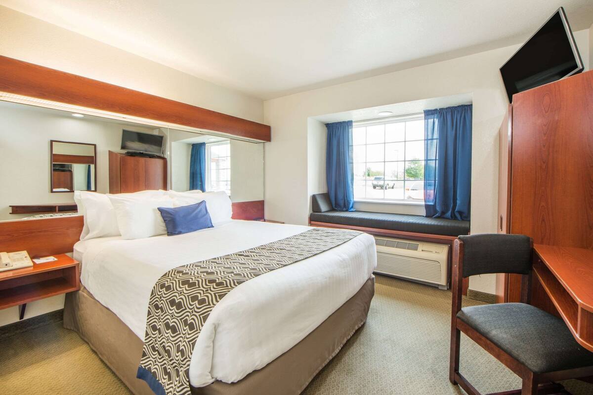 A hotel room with a large bed, a desk, a chair, a TV, a window with curtains, and a mirror.