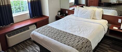 A hotel room with a large bed, a desk, a chair, a heater, a window with curtains, and a television on the wall.