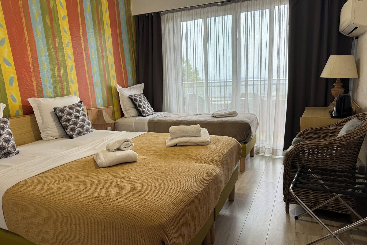 A hotel room with two beds, a wicker chair, a lamp, and a view of the outdoors through large windows.