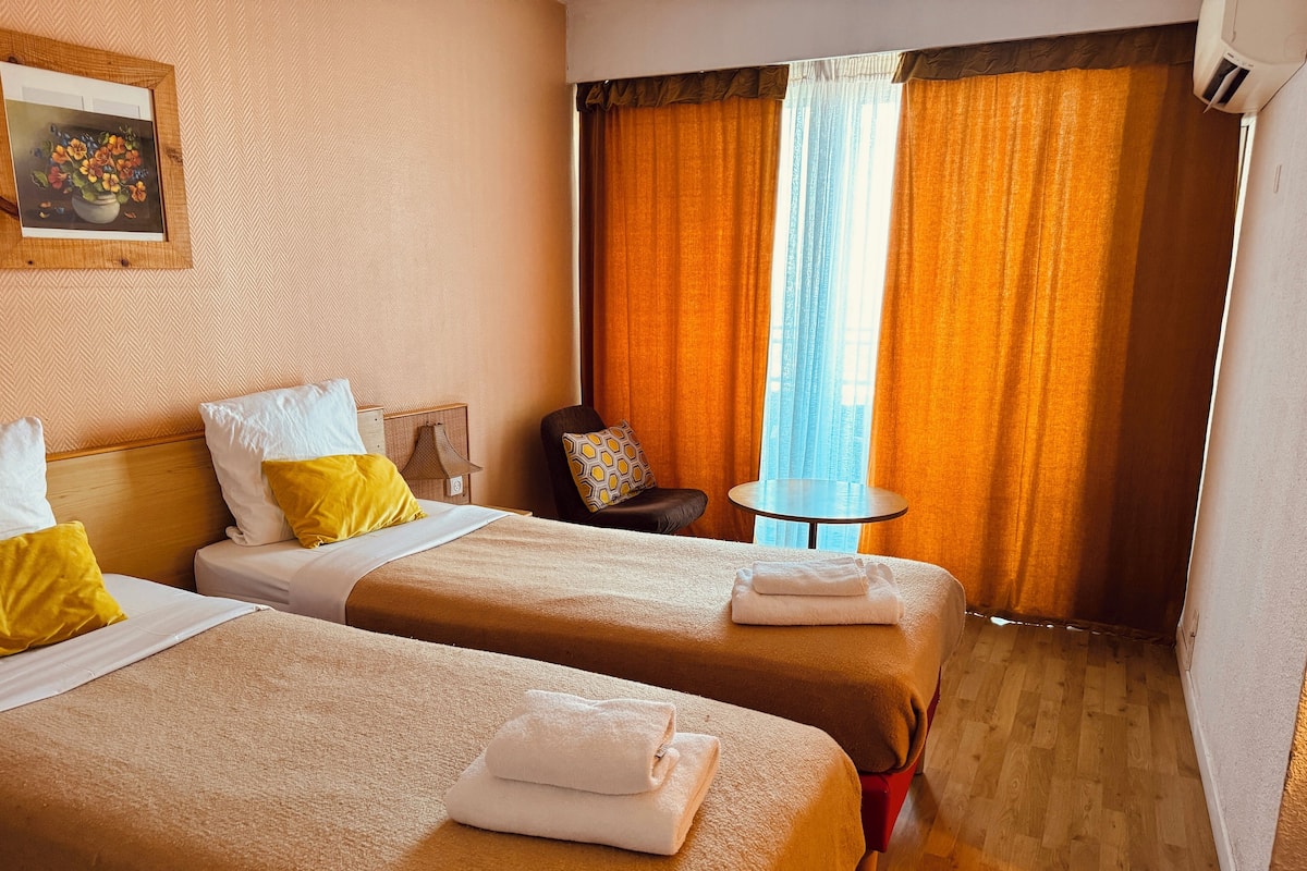 A hotel room with two beds, a small table, a chair, and a painting on the wall.