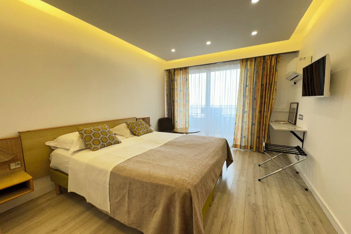 A modern hotel room with a large bed, a bedside table, a chair, a TV mounted on the wall, and a window with curtains.