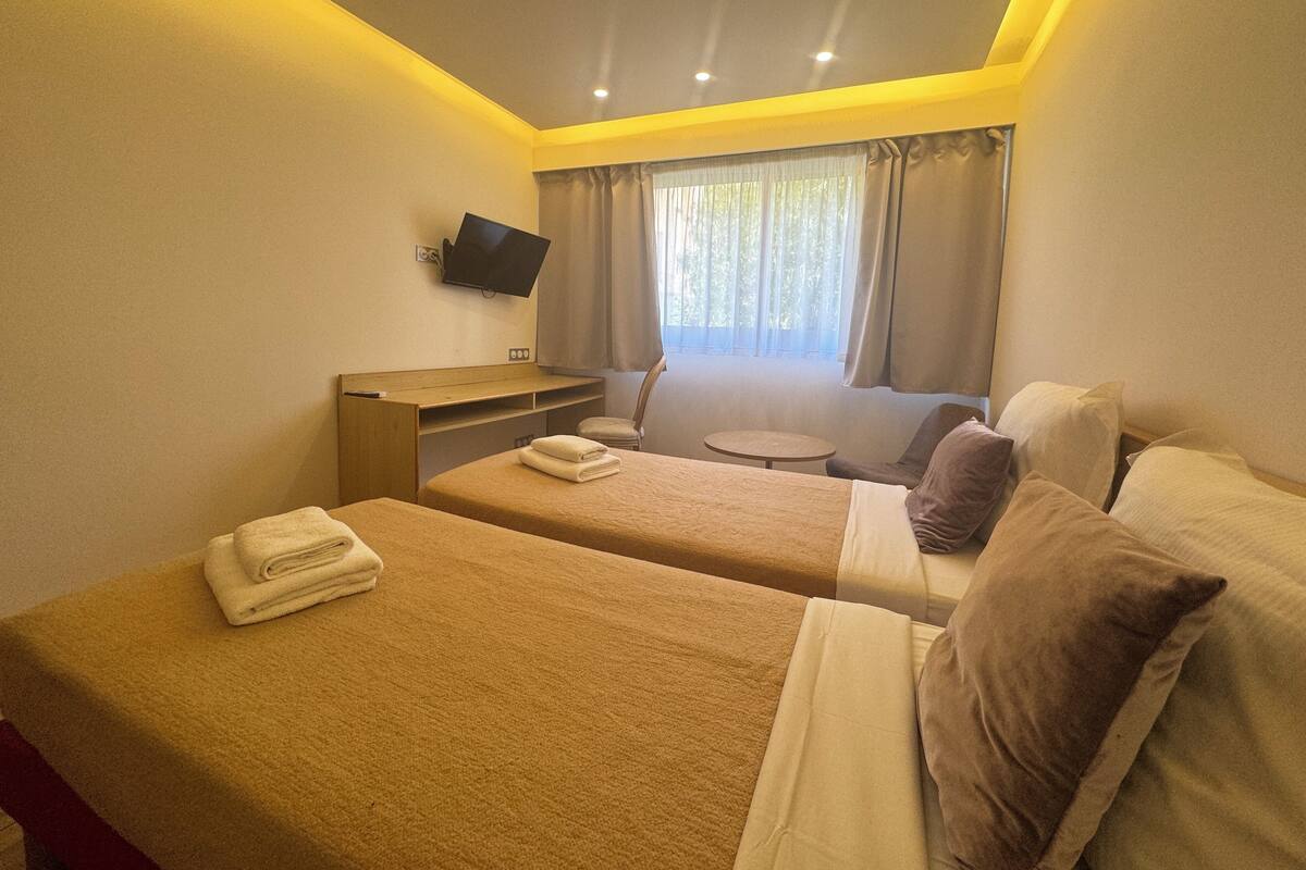 A hotel room with two beds, a desk, a TV, and a window with curtains.