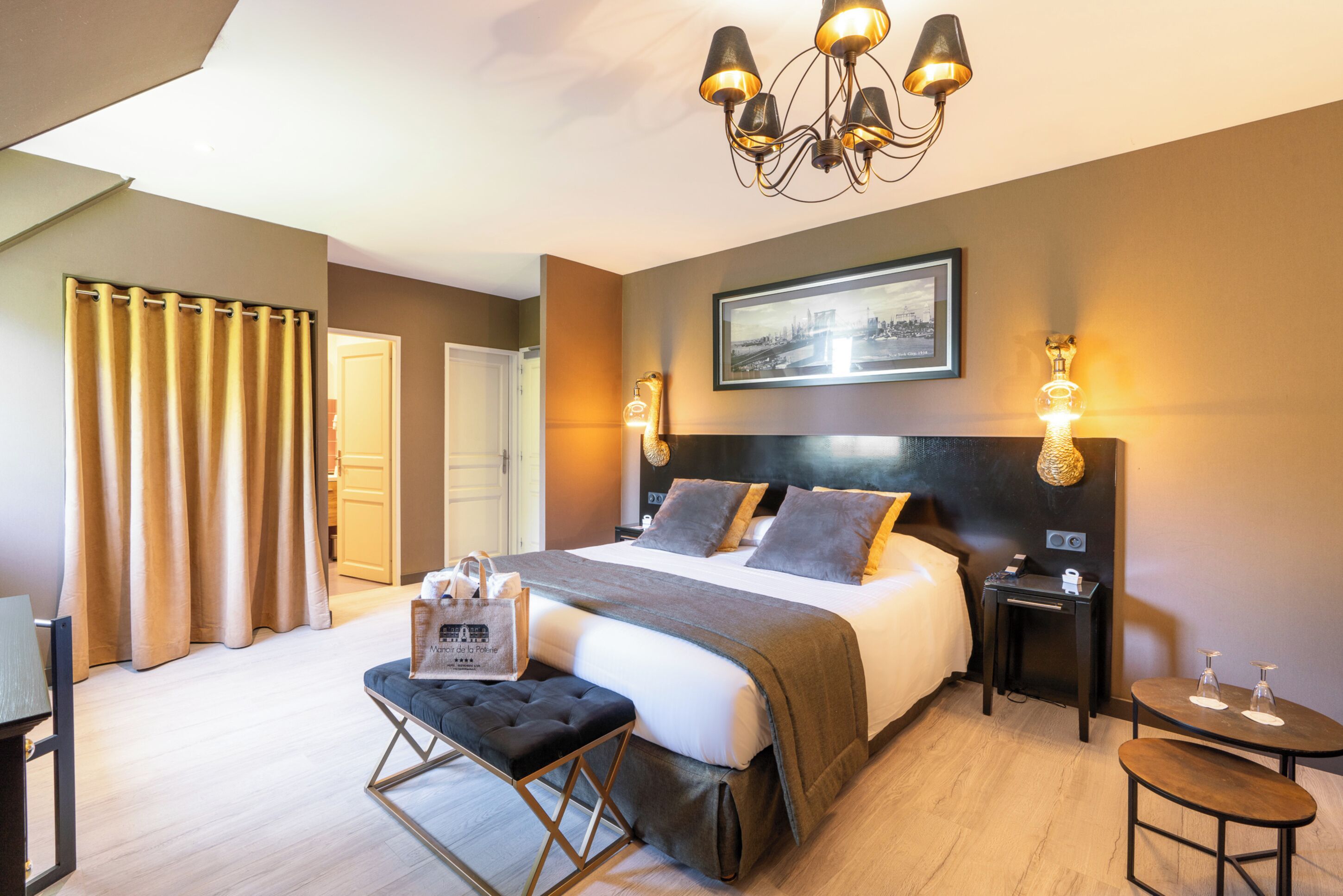A hotel room with a large bed, a bench, a small table, a nightstand, and a chandelier.