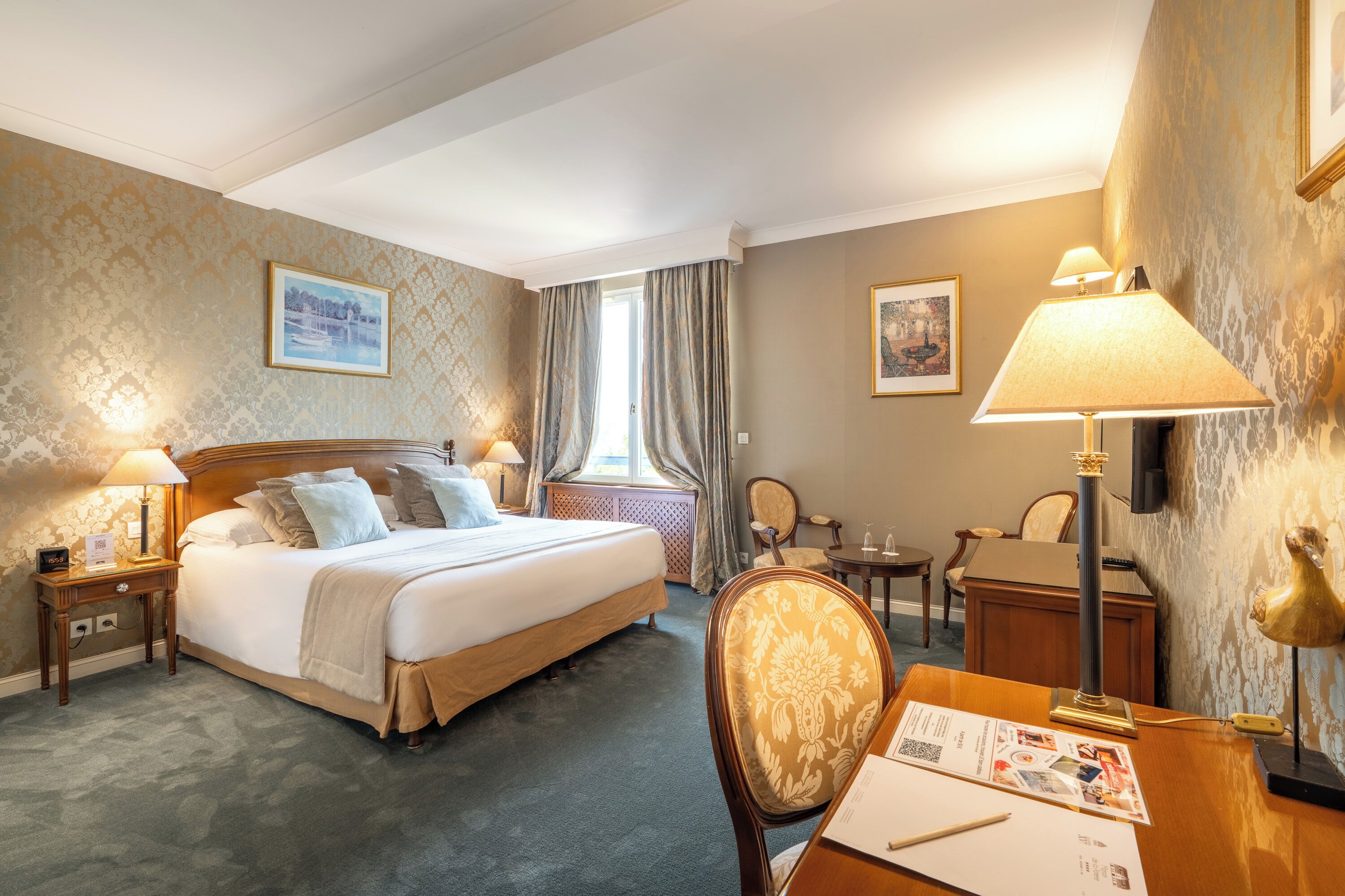 Habitación de hotel con una cama grande, dos mesitas de noche con lámparas, un escritorio con una silla, una pequeña mesa y una ventana con cortinas.