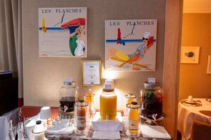 A hotel room with artwork depicting beach scenes, a minibar with various beverages, and neatly arranged dining tables.