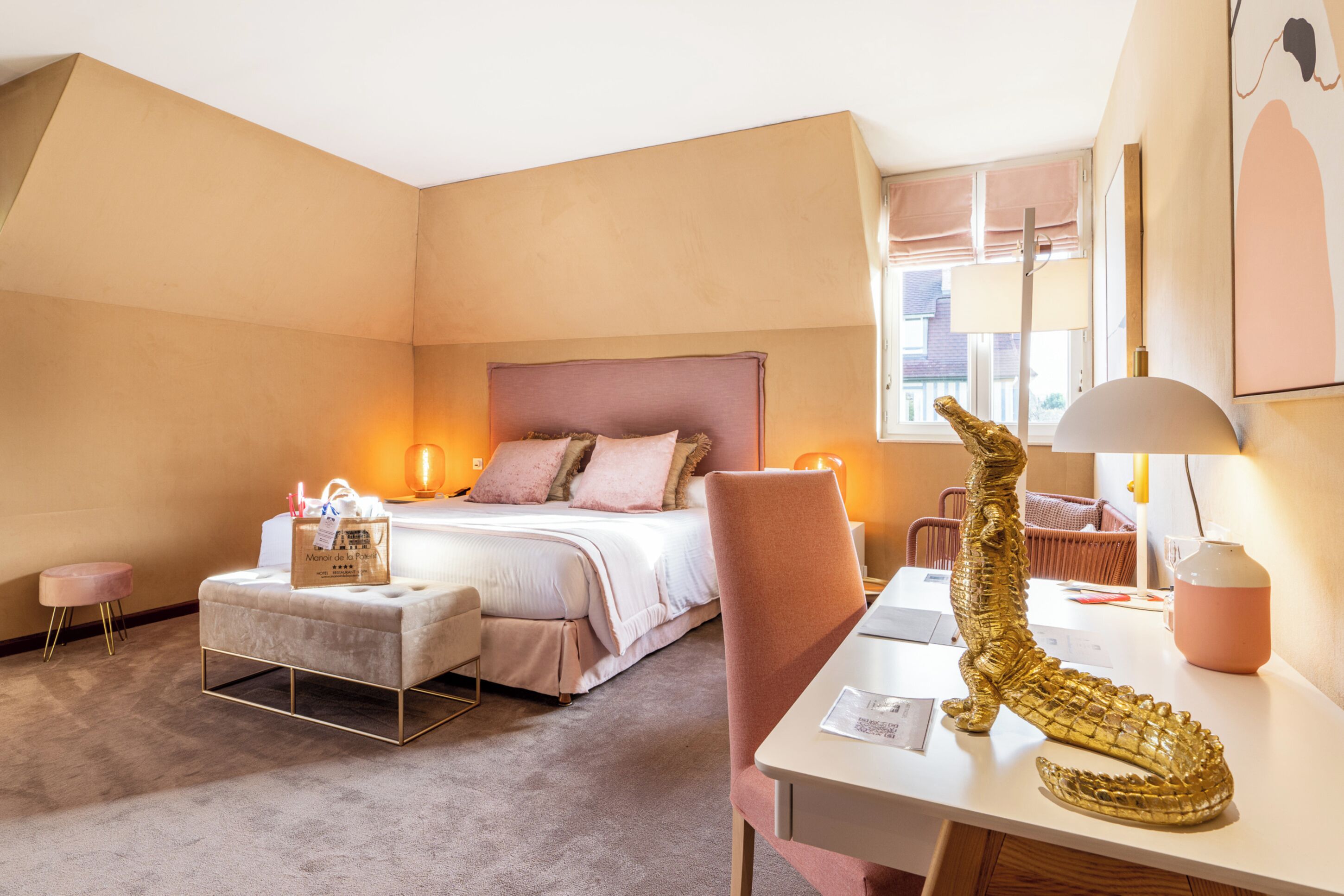 Habitación de hotel con cama, escritorio, silla, pequeño otoman, lámpara y una escultura dorada decorativa.