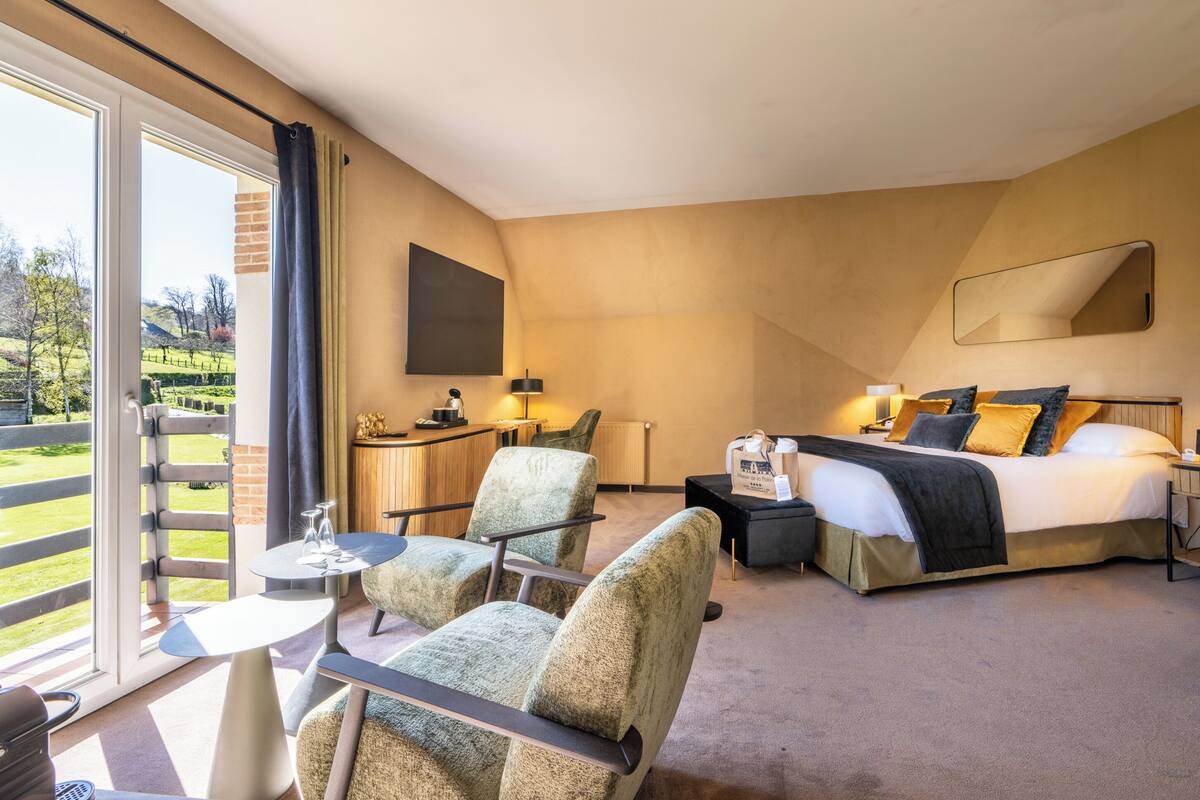 Une chambre d’hôtel comprenant un lit, deux fauteuils, une petite table, un bureau et une grande fenêtre donnant sur un jardin.