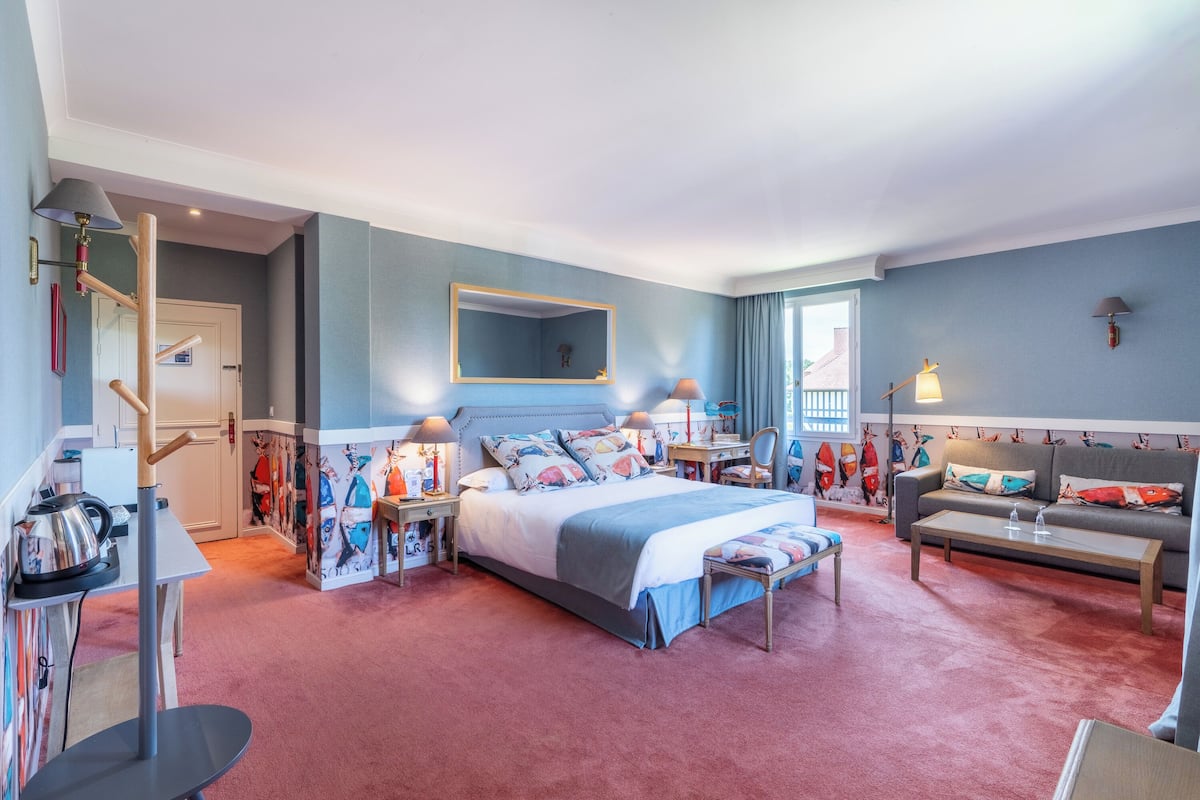 Une chambre spacieuse avec un grand lit, un canapé, un bureau et une chaise. Il y a une fenêtre avec des rideaux, un miroir et une lampe sur le bureau.