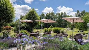 An outdoor seating area with wicker chairs and umbrellas, surrounded by lush greenery and flowering plants.