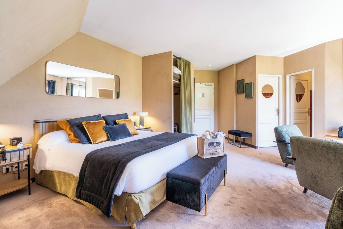 Une chambre d’hôtel avec un grand lit, un banc, une chaise, une table de chevet, un miroir et une porte donnant sur une salle de bain.