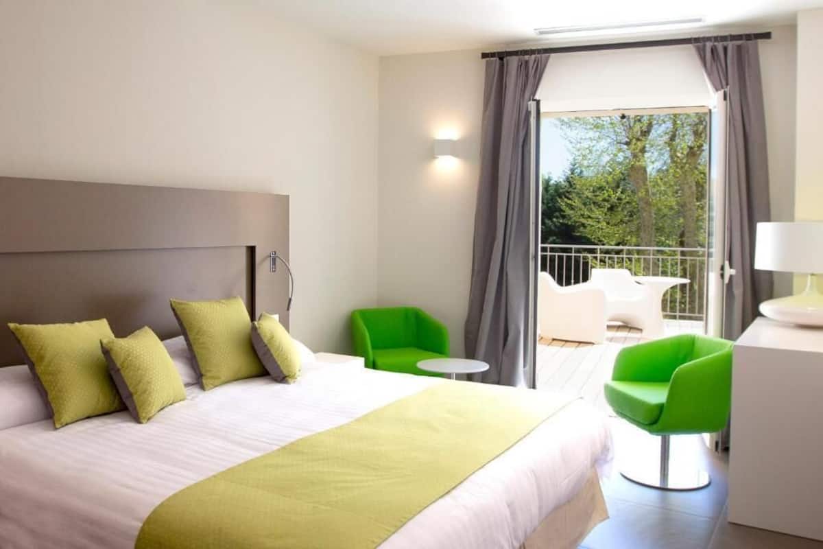 Ein Hotelzimmer mit Bett, grünen Stühlen, einem kleinen Tisch und einem Balkon mit Blick auf Bäume.