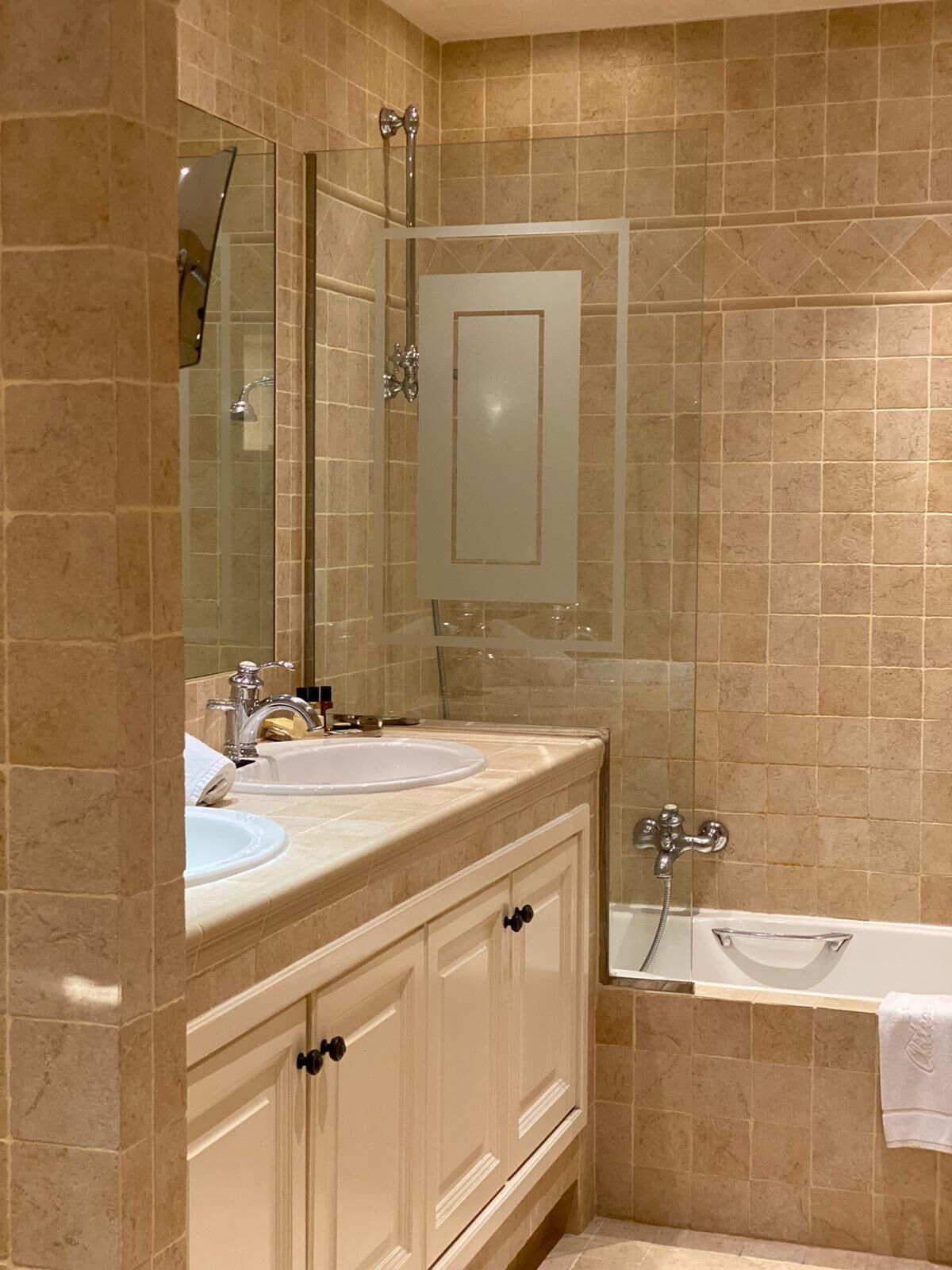 Ein Badezimmer mit beige gefliesten Wänden, einem doppelten Waschbecken, einem Spiegel und einer Badewanne.