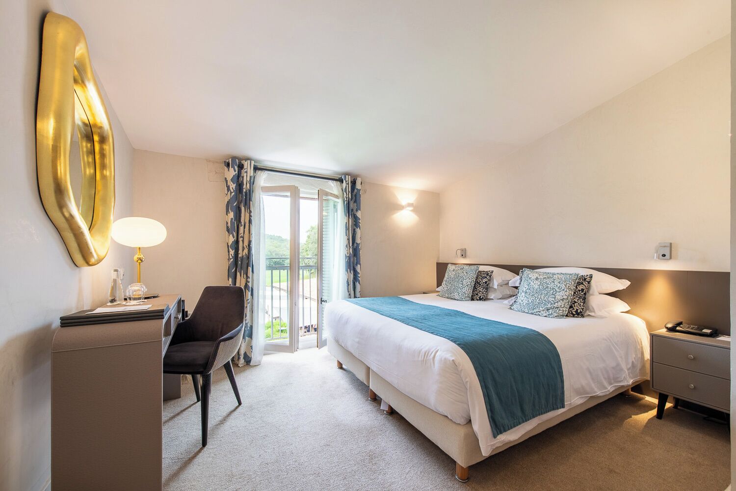 A hotel room with a bed, bedside table, lamp, chair, and a desk with a lamp.