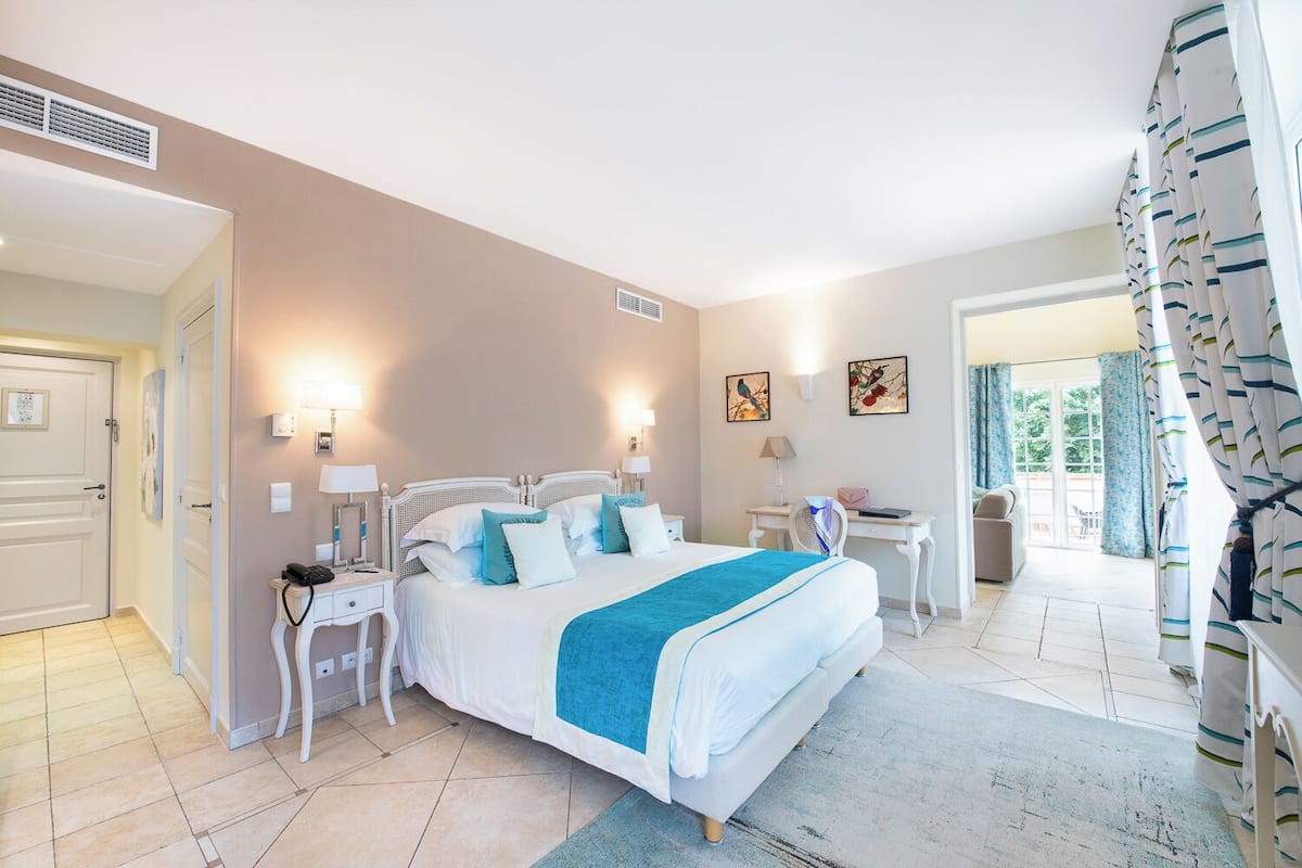 Ein Schlafzimmer mit einem großen Bett, Nachttischen, einem Schreibtisch und einem Sessel. Zwei Wandlampen und ein Fenster mit Vorhängen sind vorhanden.